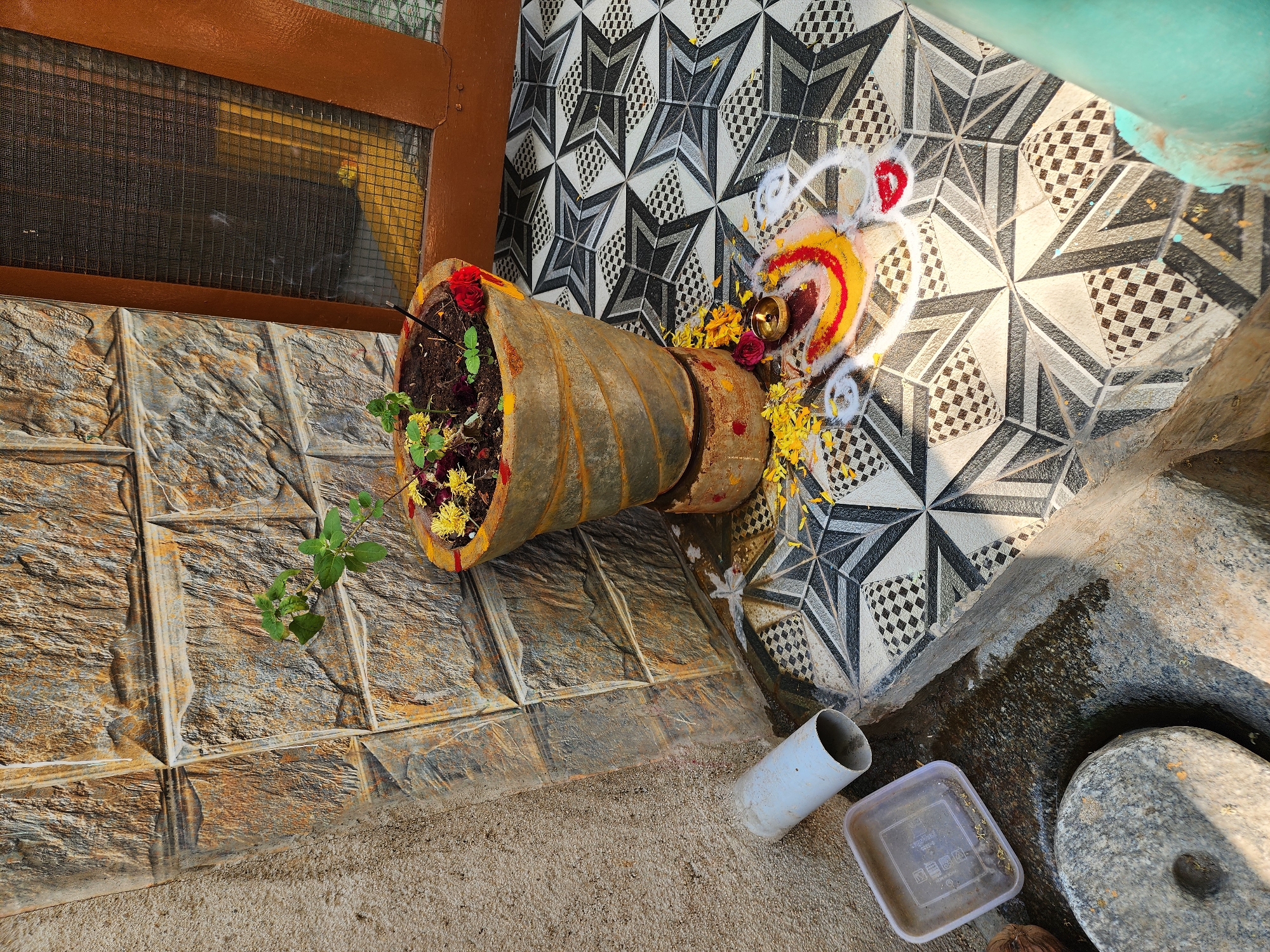 The image displays an outdoor or semi-outdoor domestic area in Pedda Kammavaripalli, India, under direct daylight. A grey, conical, ribbed plant pot, containing soil, a small green plant, and scattered yellow and red flower petals, is centrally positioned on a patterned tiled floor. The pot rests on a smaller, cylindrical stand. An incense stick protrudes from the soil. On the tiled floor, a multi-colored Kolam (Rangoli) design, created with white, yellow, and red powder, surrounds a small, lit brass oil lamp. Yellow flower petals are scattered around the lamp and Kolam. The tiled floor features a black, white, and grey geometric star-like pattern. To the right, a dark brown wooden frame with a mesh screen is visible, suggesting a door or window. Behind the plant pot, the wall is covered with rough-textured, rectangular stone-patterned tiles in shades of brown and grey. To the left, the tiled area transitions to a coarser, light brown stucco wall and a darker, rough ground surface. On this rough ground, a short section of a white PVC pipe lies horizontally, beside a translucent square plastic container. A circular grey stone object with a central bore hole is partially visible in the lower left corner. A vertical greenish-blue object is partially visible on the far right edge of the frame. No individuals are present.