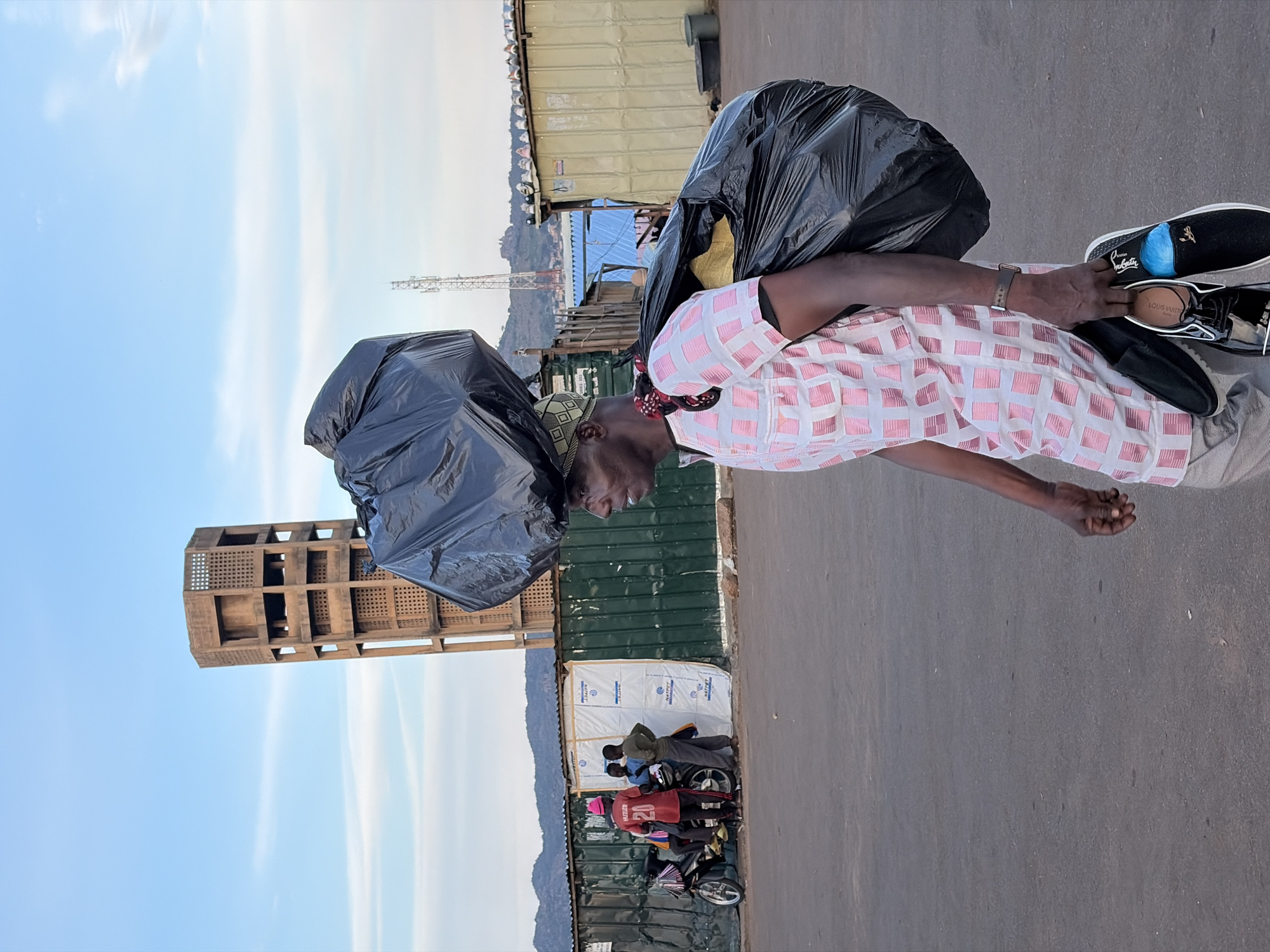 A medium shot captures a side profile of a person walking in Jos, Nigeria. The individual is carrying a large, black garbage bag balanced on their head and another large, black bag slung over their right shoulder. They are wearing a pink and white checkered short-sleeved shirt and light-colored pants, and are holding two pairs of shoes in their right hand. In the background, several people are gathered around a motorcycle near a green corrugated metal fence. Behind the fence, a tall, Brutalist-style concrete tower stands prominently against a clear blue sky with some wispy clouds. Other structures, including a telecommunications tower and what appear to be small buildings or stalls, are visible further in the background, with mountains in the distance. The ground is paved with asphalt.