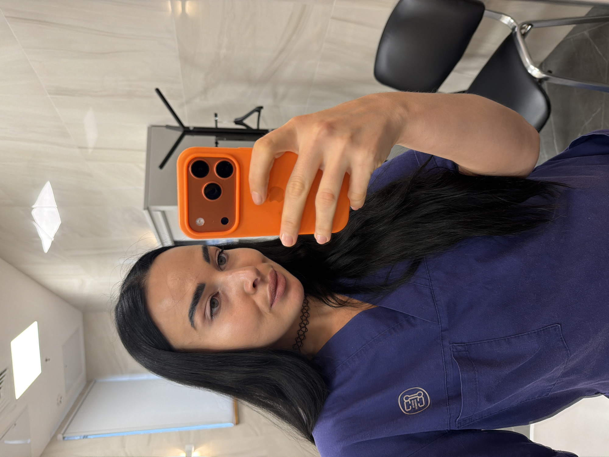 A woman takes a mirror selfie in what appears to be a clinic or professional office in Saint Petersburg, Russia. She holds an orange-cased smartphone, likely an iPhone, with her right hand, positioning it to capture her reflection. Her long, dark hair cascades over her left shoulder and chest. She is wearing a dark blue scrub top, which features a silver-colored logo on the left chest area. The logo is mirrored due to the selfie and reads "LAMBERGER SCRUBS &" with additional, unclear text below it. She has a neutral to slightly pouty expression, with defined eyebrows and a nude lip color. A black choker necklace with a patterned design is visible around her neck.
The background reveals a clean, modern interior. Behind her, there's a light-colored, possibly tiled, wall and a dark office chair with a chrome frame. Another dark chair is partially visible further back. To her left, bright square light fixtures are mounted on the ceiling or wall, illuminating the room. A white surface, possibly a whiteboard or counter, is also visible on the left side of the frame. The overall impression is a professional and well-lit environment.