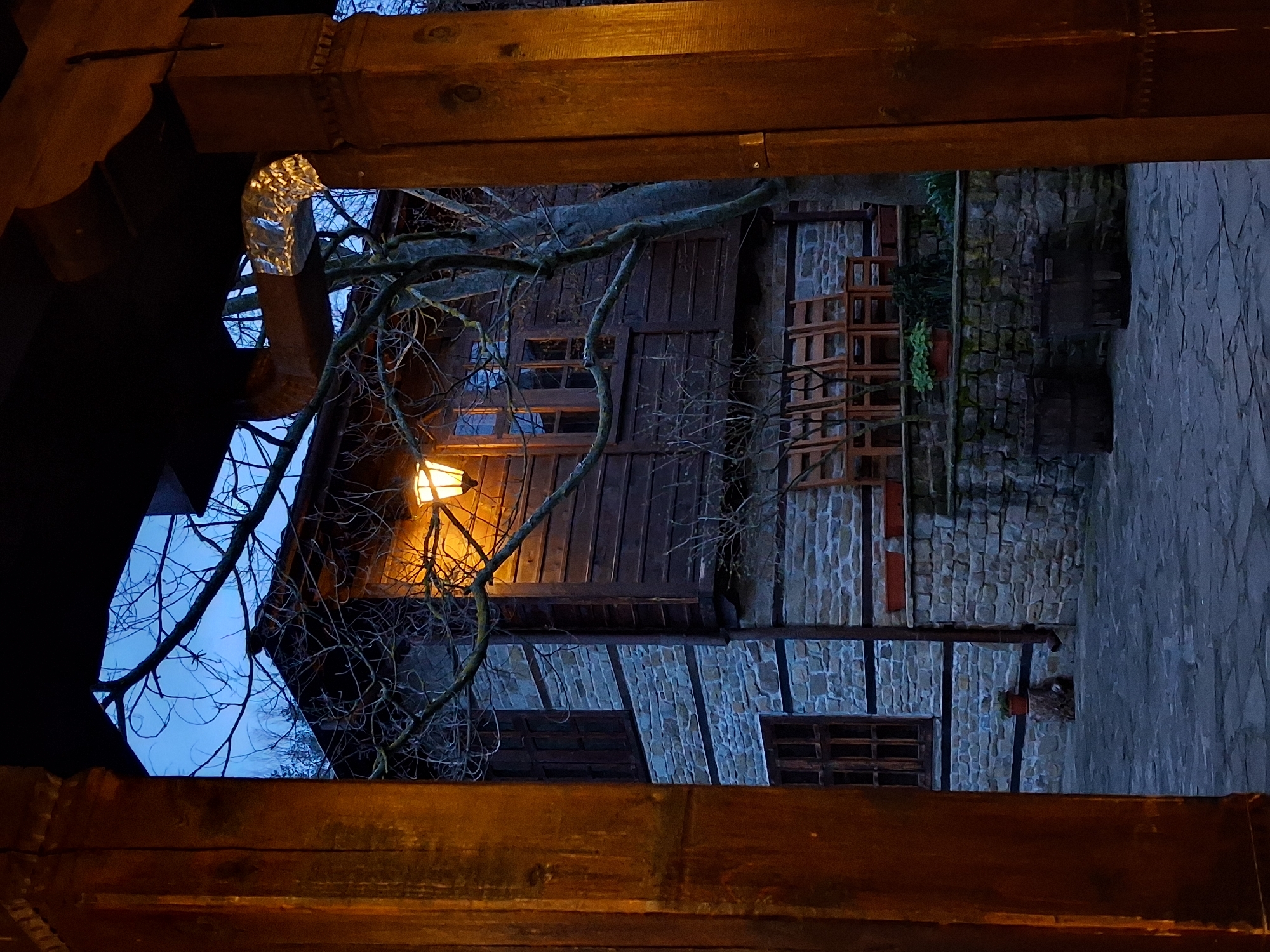 The image shows a building in Katunishte, Bulgaria, framed by dark wooden beams, possibly part of a structure like a porch or window frame. The building appears to be old, with stone and timber framing, and a dark wooden facade. A lit lantern is visible near the building, and a bare tree branch is in the foreground. A stone wall is visible to the right. The overall lighting is dim, with a focus on the lantern's glow.