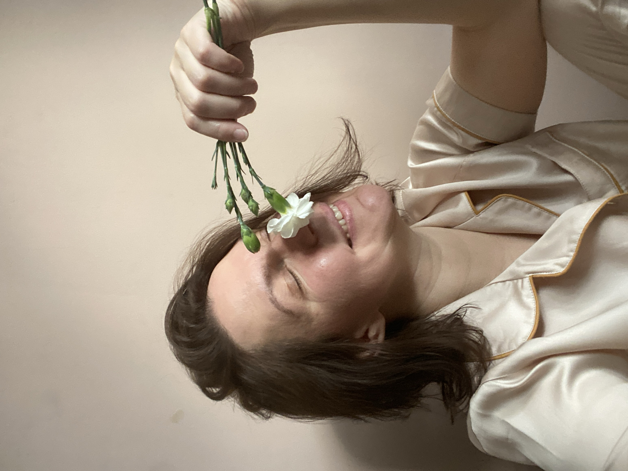 This close-up, rotated image, potentially taken in Chaikovsky, Russia, features a woman interacting playfully with flowers. The woman, appearing from her forehead to her chin, has short to medium-length dark hair and is captured in profile, looking upwards with her eyes closed and a wide, joyful smile. Her right arm and hand are raised, holding a small bunch of green-stemmed flowers. One small, white flower is positioned delicately near her nose, while several other flower buds remain closed on their stems.
She is dressed in light beige or champagne-colored pajamas or loungewear, which appears to be made of a smooth, slightly shiny fabric like satin or silk, accented with a darker trim along the seams and collar. The background is a plain, light beige wall, which provides a simple and uncluttered setting, allowing the viewer's focus to remain entirely on the woman's expressive face and the flowers. The overall impression is one of happiness, contentment, and a moment of simple pleasure or sensory appreciation. There is no visible text in the image.