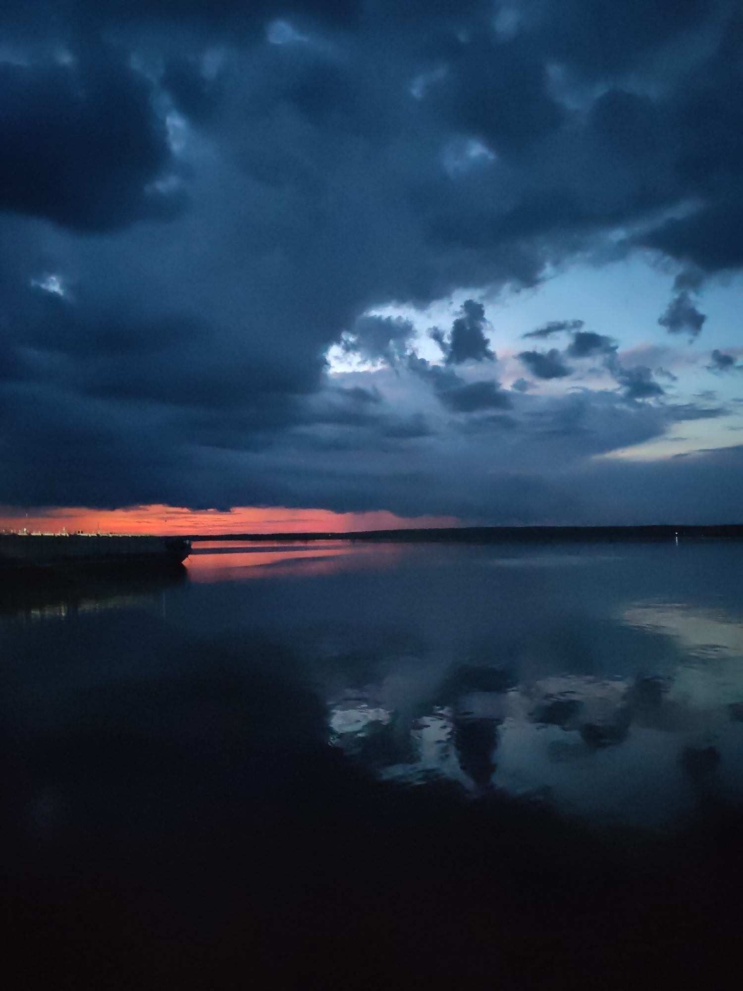 An image captured at dusk in Rybinsk, Russia, shows a body of water with a striking sunset in the background. Dark, ominous clouds dominate the sky, with breaks of lighter blue and hints of pink and orange from the setting sun visible near the horizon. The water reflects the dramatic sky, creating a mirror-like effect. A dark silhouette of a large boat or barge is visible on the left side of the frame, partially submerged in the water. The overall scene evokes a sense of brooding beauty and the approaching night.