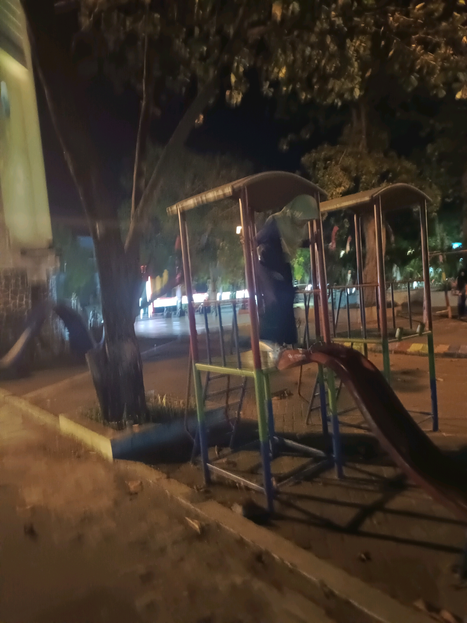This is a night-time photograph of a children's playground, possibly located in Lamongan, Indonesia. A metal playground structure with red, green, and blue supports is the central focus. A dark slide curves downwards from the structure. In the background, blurred trees and lights suggest an urban or park setting. The ground appears to be paved or dirt. A person wearing dark clothing and a head covering is visible within the playground structure. The overall image quality is low, with blur and low light conditions.
