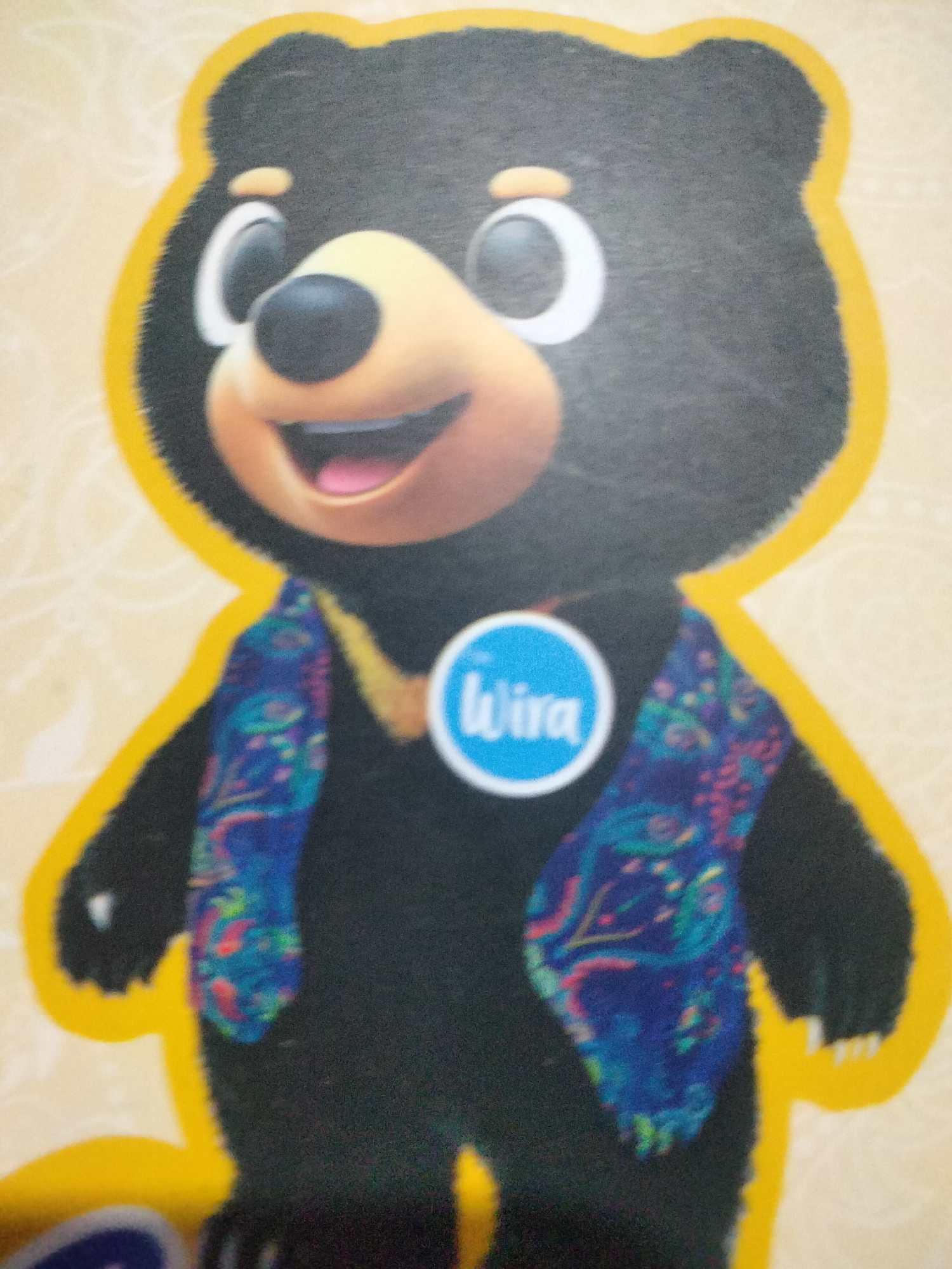 A cartoon black bear character wearing a blue and colorful patterned vest stands against a beige background with a subtle patterned texture. The bear has a round, yellow outline and a large, smiling face with wide, black eyes with white highlights, and a pink tongue visible. A blue circular badge with the word "Wira" in white text is prominently displayed on its chest. The bear's fur appears soft and fluffy. The image is taken from a frontal perspective, with the bear occupying the majority of the frame. There are no discernable elements within the image that would indicate a specific location in the city of Payagaboh, Indonesia.