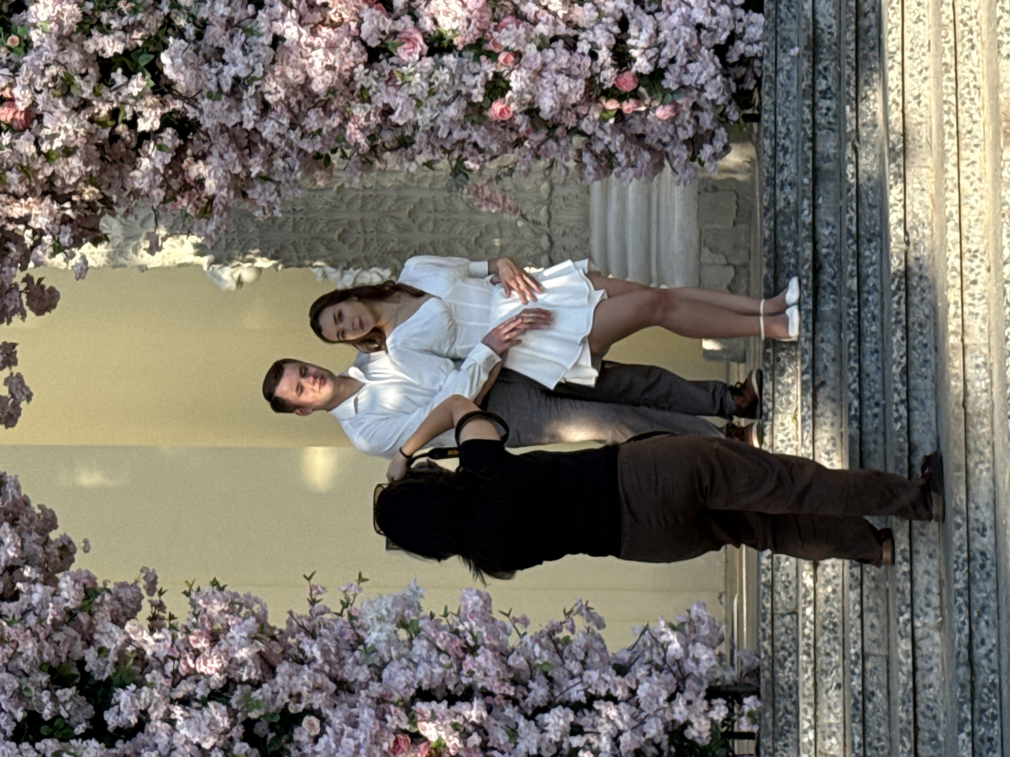 In front of a classical building with a yellow facade, a young couple poses on a stone staircase adorned with abundant pink floral arrangements. The man, wearing a white collared shirt and grey trousers, stands to the left with his arm around the woman. The woman, in a short white dress, looks towards the camera with her hand on the man's chest and the other hand on her hip. To the left of the couple, a woman with dark hair, wearing a black t-shirt and brown pants, is taking a photograph of them with a camera. The stairs are wide and made of stone, with visible texture and wear. The scene is illuminated by natural light, casting shadows on the steps and highlighting the textures of the flowers and architecture. The architecture suggests a public building, possibly a museum or historic landmark, in Moscow, Russia.