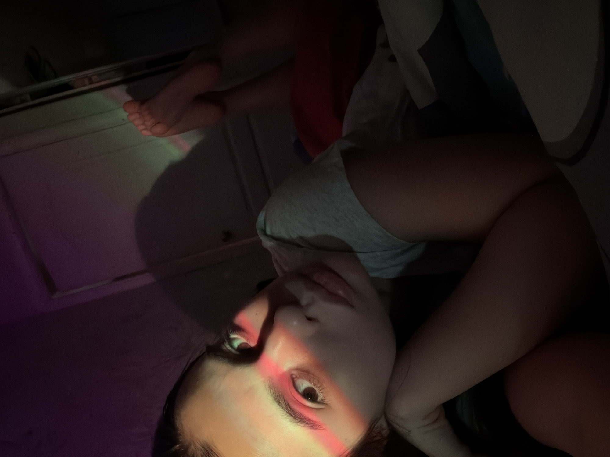 A rotated image captures a young individual, likely female, in a dimly lit indoor setting in Chaikovsky, Russia. The person is reclining or lying down, with their head positioned towards the bottom right of the frame and bare feet visible in the upper left, resting on what appears to be a white cabinet or piece of furniture. They are wearing a light-colored top, possibly grey.
The scene is dramatically lit by strong, colored light beams. A prominent red beam cuts across the person's face, highlighting their right eye, nose, and mouth, while also casting a glow on their left eye area. Fainter green and purple hues are also visible in the background, particularly on the surface where the feet are resting. The person's expression is neutral and calm, with a slight pout to the lips. The overall darkness of the background suggests the image was taken at night or in a very dim room, emphasizing the artificial light. No visible text is present in the image.