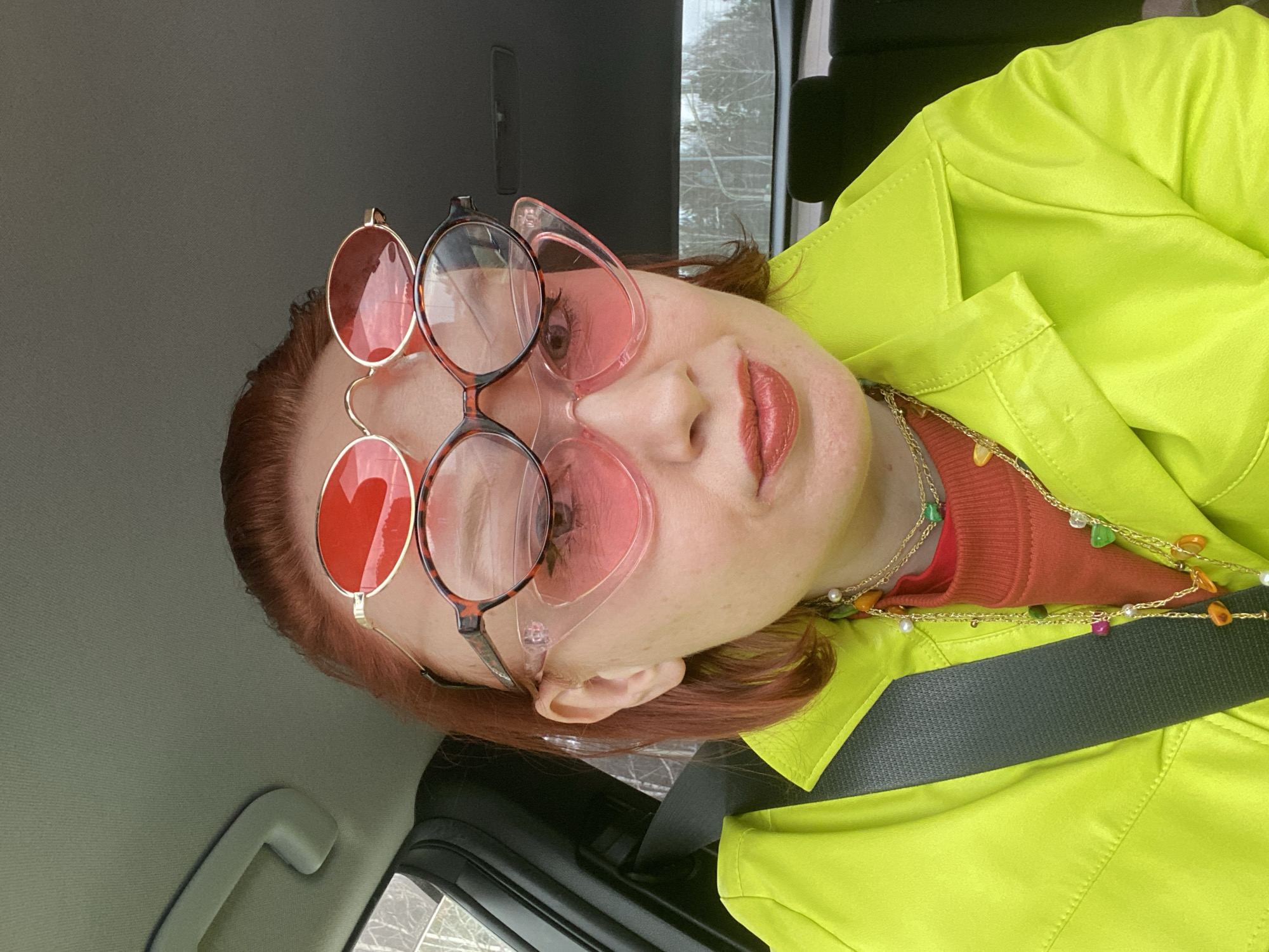 The image depicts a person inside a vehicle, likely a car, wearing three pairs of glasses stacked on their face. The top pair is round with red lenses, the middle pair is round with a tortoiseshell frame, and the bottom pair is a pink, heart-shaped frame. The person wears a bright yellow jacket over a reddish-brown top and several layered necklaces with colorful beads. A seatbelt crosses their body. Through the car window, trees and snow are visible, suggesting a cold climate. The interior of the car is a standard car interior. There are no explicit indications this is the city of Yekaterinburg, Russia.