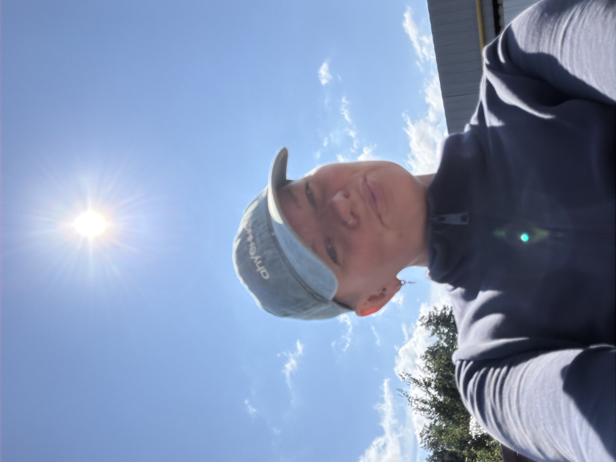 A person wearing a blue baseball cap and a dark zip-up sweatshirt is taking a selfie outdoors. The sun is bright and high in the sky, casting rays of light. There are some clouds in the blue sky, and trees and a building are visible in the background. The image appears to be taken in Chaikovsky, Russia.