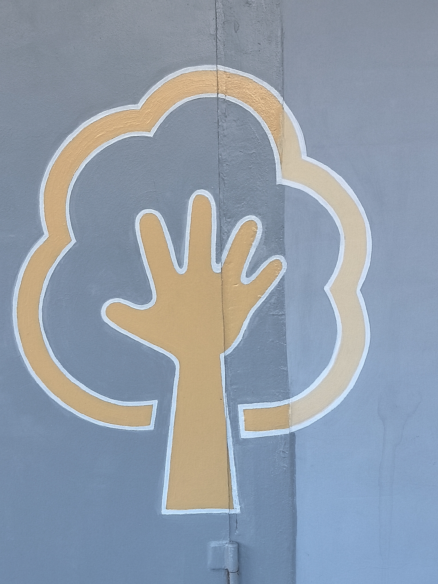 The image displays a painted mural on a blue-gray wall in Sarapul, Russia. The mural depicts a stylized tree, rendered in orange-gold with white outlines. A human hand, palm facing forward with fingers spread and upward, forms the tree's trunk and lower structure. Above the hand, a series of contiguous, cloud-like rounded shapes complete the tree's canopy. The wall surface is textured and painted, featuring a distinct vertical seam or structural joint located approximately one-third from the right side of the frame. Subtle surface imperfections and variations in paint application are discernible across the blue-gray background.