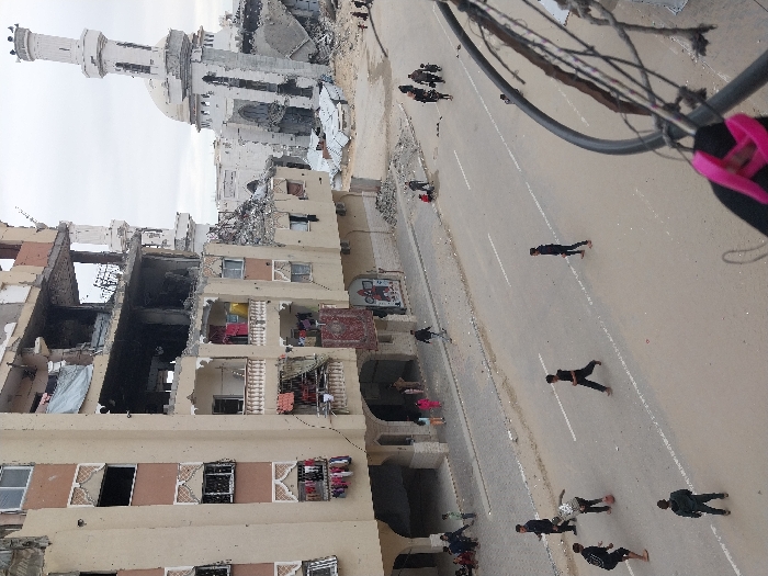 The image depicts a street scene in Khan Yunis, Palestinian Territory, characterized by extensive structural damage to buildings under an overcast sky.
In the foreground and midground, a paved road with white lane markings spans horizontally. On this road, a group of five young males are playing soccer with a red ball. Another male figure walks further down the street to the right.
To the left of the road, a multi-story building, likely an apartment complex, stands with significant damage. Its upper floors are partially collapsed, exposing interior rooms and structural elements. Balconies on the intact lower-middle floors display clotheslines with hanging laundry. Several individuals, including adults and children, are present on the sidewalk beneath this building.
In the background and to the right, a large mosque is severely damaged. Its dome is partially destroyed, and one minaret is heavily fractured and leaning, while the other appears to be a partial ruin. Piles of rubble surround the mosque. Near the mosque ruins, a small group of people, including adults and children, are walking.
The lower right foreground is partially obstructed by a dark, curved object, likely an overhead cable or pipe, from which a pink strap hangs. Debris and rubble are visible along the road edges and near damaged structures.