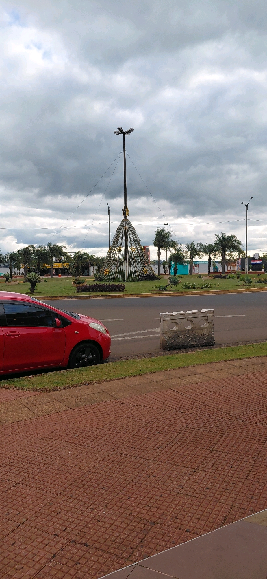 The image depicts a street scene in Encarnación, Paraguay, with a large, conical Christmas-themed light display as the central focus. The display is elevated on a tall pole and adorned with golden stars. Palm trees and other greenery surround the display, suggesting a tropical or subtropical climate. In the foreground, a red car is parked on the side of the road, next to a sidewalk paved with reddish-brown interlocking tiles. A concrete barrier with circular cutouts and arrow-like patterns is positioned between the road and the sidewalk. The sky is overcast with dark, heavy clouds, indicating potentially inclement weather. The overall atmosphere is that of an urban public space, possibly a park or a roundabout, decorated for the holiday season.