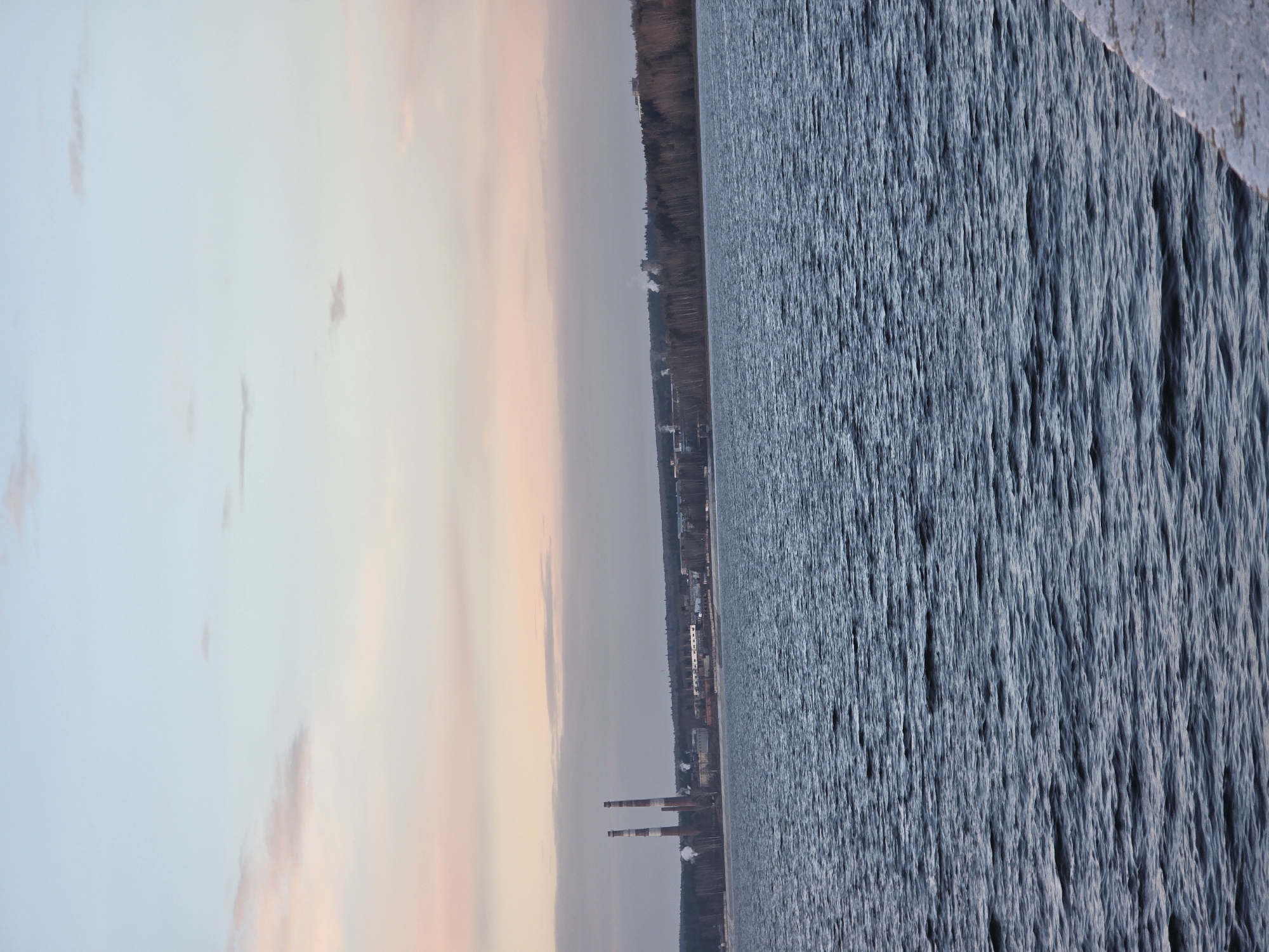 The image depicts a body of water with slight ripples, likely a lake or reservoir, adjacent to the city of Chaikovsky, Russia. The shoreline is visible, with a tree line and buildings in the distance, including industrial structures with smokestacks emitting smoke. The sky is overcast, with a hint of pastel colors near the horizon, possibly indicating sunrise or sunset.