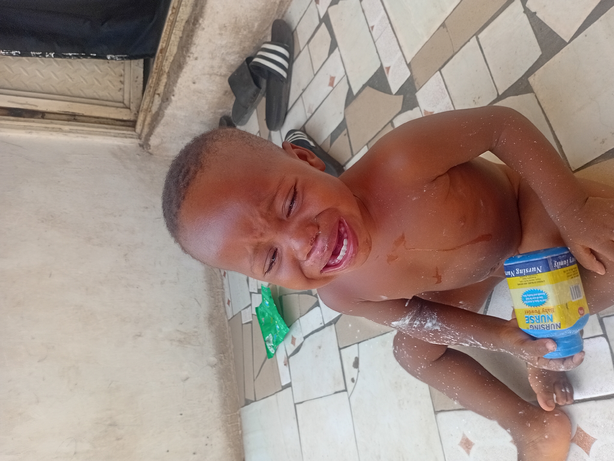 An overhead shot captures a young child, appearing to be a toddler, in a state of distress, crying with their mouth open and eyes closed, while sitting on a tiled floor. The child has dark skin and appears to be naked or partially clothed, with a white, powdery substance (likely baby powder) liberally sprinkled across their body, especially on their arms, chest, and legs. Some powder is also visible on the floor around them.
The child is holding a blue and yellow container of baby powder in their left hand. Visible text on the container includes "NURSING NURSE Baby Powder" and "A PRODUCT OF PLASCO INDUSTRIES LTD, LAGOS, NIGERIA". Other text like "family" and "NURSING Mam" is also partially discernible.
The setting appears to be an outdoor or semi-outdoor area, possibly a patio, porch, or a simple bathroom space, given the visible concrete and tiled floor. The floor is covered with beige and white rectangular tiles, some with small reddish-brown accents, and appears somewhat worn or dirty in places. In the background, to the left of the child, a pair of black slides with white stripes is visible. Further in the background, a door frame or threshold with a metallic, textured surface can be seen. A crumpled green object, possibly a wrapper or plastic, lies on the floor near the child's head. The scene is illuminated by daylight. The child's intense crying is the central focus, indicating an upset or uncomfortable moment, possibly related to the powder.