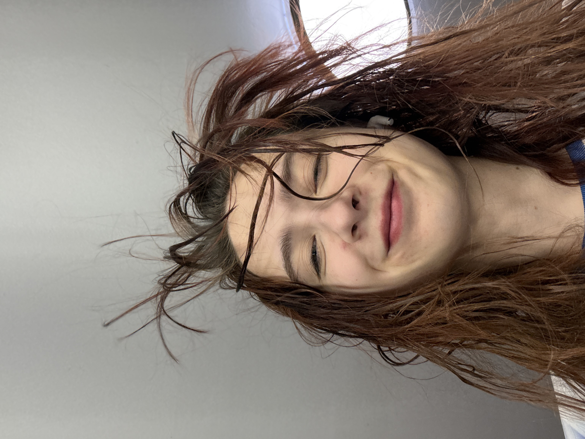 A close-up, tilted portrait depicts a person with wet, brown hair. One ear is visible, containing a white earbud. The person is smiling with their eyes partially closed. The background appears to be a plain gray wall. The location in the city of Прикамский, Russia is not evident.