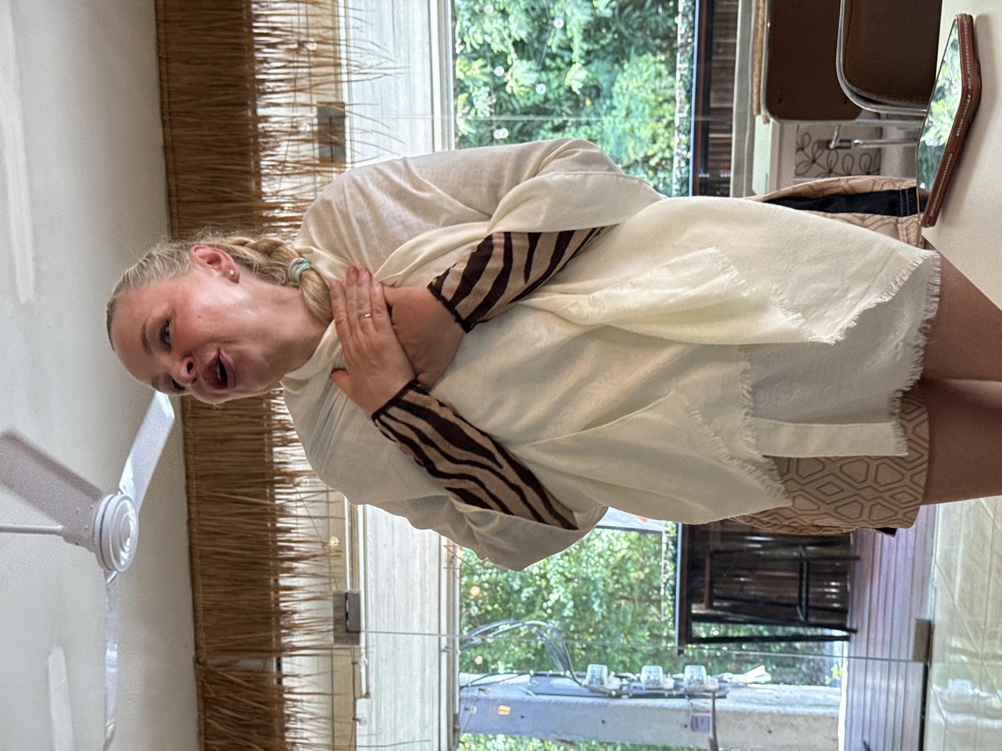 A woman stands indoors in what appears to be a resort or restaurant with a thatched roof ceiling and a ceiling fan. She is wearing a white shawl draped over her shoulders, a white long-sleeved shirt with striped cuffs, and a patterned skirt. Her hands are clasped over her chest, and she has an expressive look on her face, with her mouth open. The background shows a large window overlooking lush greenery, suggesting an outdoor or garden setting. A table with a smartphone on it is visible in the foreground. The scene is well-lit, with natural light coming from the window and artificial lights from the ceiling. The overall impression is that of a candid moment captured during a conversation or performance.
