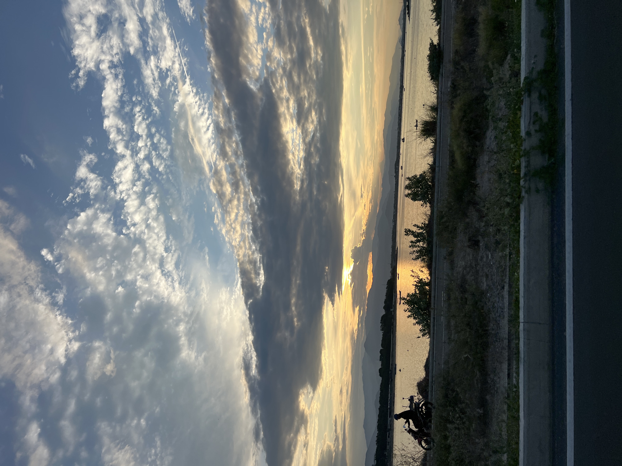This is a vertical, landscape-oriented photograph taken at sunset in Bac Cam Ranh Ward, Vietnam. The image captures a serene scene with a vast sky filled with dramatic clouds transitioning from bright white and yellow near the horizon to darker grays and blues higher up. A contrail from an aircraft is visible in the upper left portion of the sky.
The horizon line is dominated by a body of water, likely a bay or gulf, reflecting the warm hues of the setting sun. Distant mountains are silhouetted against the bright sky. Scattered on the water are several small boats.
In the foreground, a paved road runs along the edge of the water. To the left of the road, there is a strip of vegetation, including some trees. A person is silhouetted on a motorcycle parked on the side of the road, facing the water. The setting sun casts a warm glow on the scene, suggesting a peaceful and tranquil moment. The overall mood is one of calm and natural beauty. No text is visible in the image.