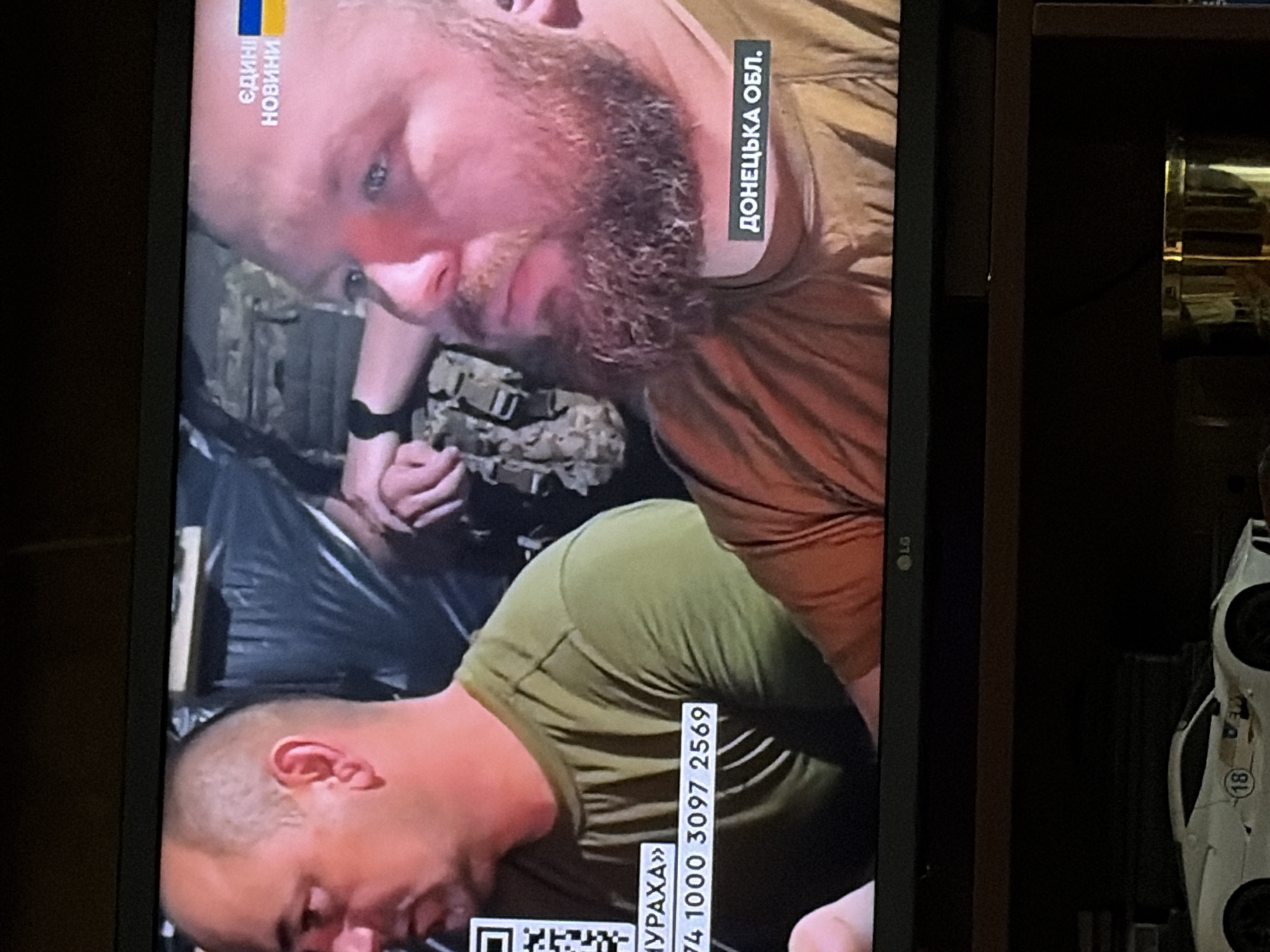 The image shows a television screen displaying a news segment. Two men are visible. The man on the left has short hair and is wearing a green t-shirt. Text overlays on his shirt include a QR code and what appears to be a phone number. The man on the right has a beard and is wearing an orange t-shirt. Ukrainian text and the Ukrainian flag are visible in the upper right corner of the screen. Text in the lower right corner reads "ДОНЕЦЬКА ОБЛ.", which translates to "Donetsk Oblast." Below the television, a white toy car with the number "18" on it is visible. The scene appears to be indoors.