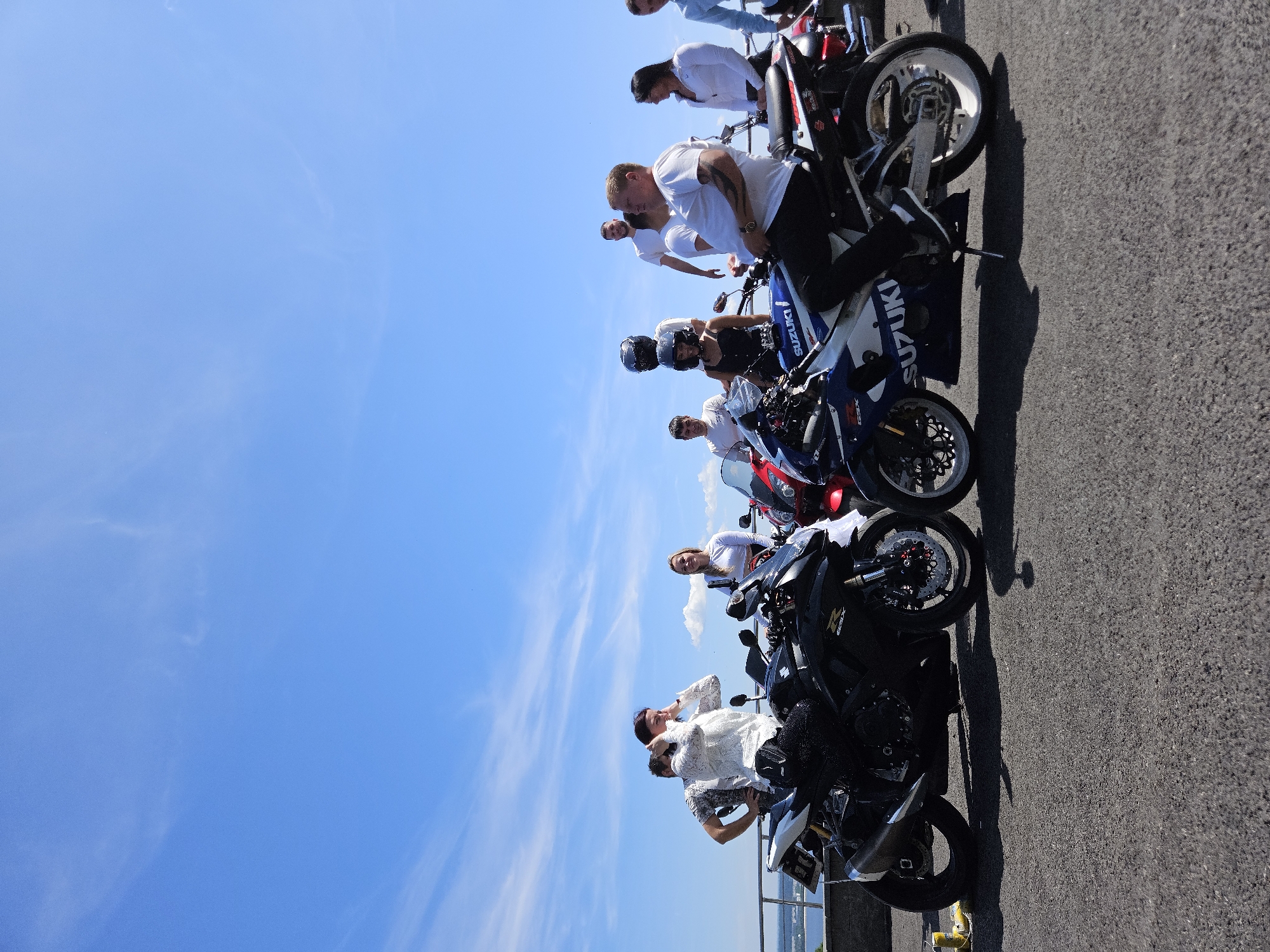 The image shows a group of people gathered around several motorcycles on a paved surface, likely a road or parking area, under a clear blue sky with wispy clouds. The scene appears to be outdoors, with a railing and a hint of water in the background on the left, suggesting a waterfront location.
In the foreground, there are two prominent sport motorcycles. The one on the left is black with silver accents, and the one on the right is white and blue with "SUZUKI" branding. Both motorcycles have their kickstands down.
Several individuals are present, some standing and some sitting on or near the motorcycles. On the left, a man in a patterned shirt is leaning on a railing. Next to him, a woman in a white lace top is adjusting her hair, with a black motorcycle in front of her. To her right, a blonde woman in a white top is standing by a motorcycle.
Further back, a man in a white t-shirt stands behind a woman wearing a black top and a helmet. Another man in a white t-shirt is visible further back. On the far right, a man in a white shirt is sitting on a motorcycle, looking to his left. Next to him, a woman in a white shirt is standing. A man in a blue shirt is partially visible on the far right.
The image captures a moment of informal gathering, possibly a motorcycle meet-up or outing. The overall atmosphere is casual and sunny. The provided context of Ulyanovsk, Russia, places this scene within that specific geographic location.