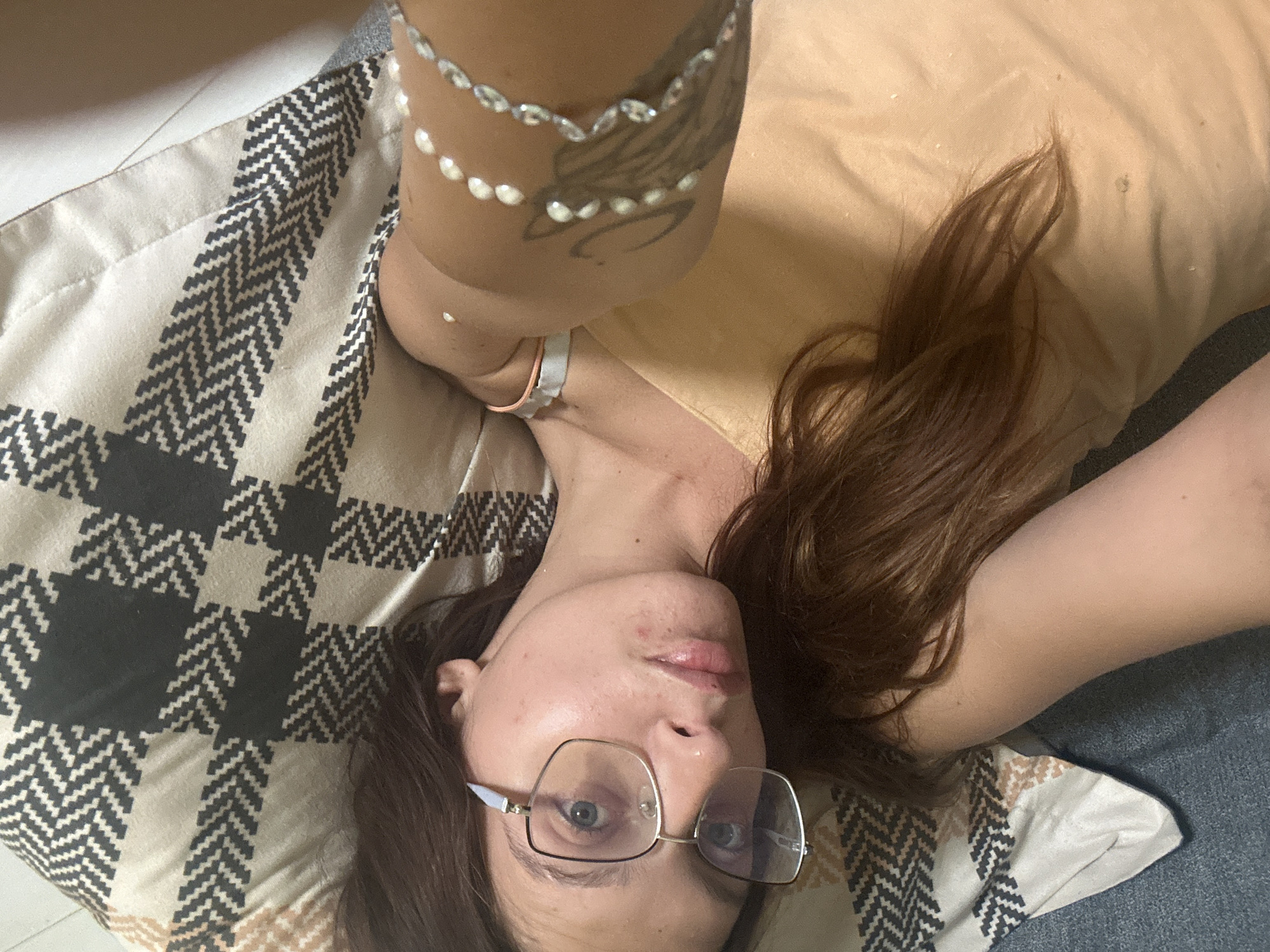 A woman with long, brown hair and blue eyes is lying on her back on a patterned blanket. She is wearing glasses and a light orange top. A bracelet made of white beads is visible on her right arm, and a tattoo is present on her left arm. The image does not provide information about a specific location in Tryavna, Bulgaria.