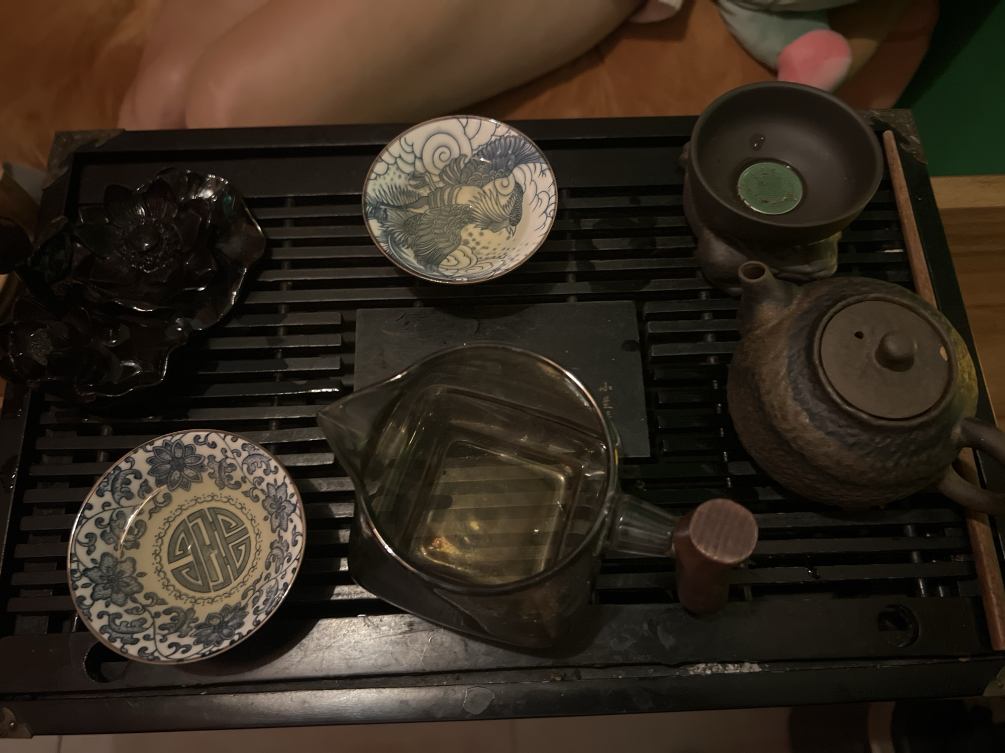 The image depicts a tea set arranged on a black, slatted tray. The set includes a dark, ornate bowl, two small decorative plates, a clear glass pitcher filled with liquid, and a rustic, brown teapot. The scene appears to be indoors. No people are actively involved in the scene. The location is Unknown, Unknown.