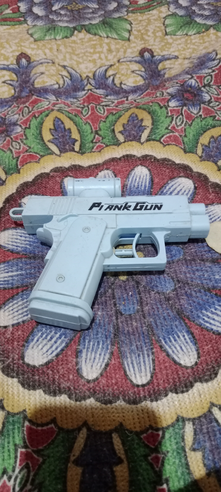 A light blue toy gun with the words "PLANK GUN" printed on its side is placed on a colorful patterned fabric. The fabric has floral motifs with intricate designs in shades of red, green, yellow, blue, and purple. The toy gun is positioned diagonally across the fabric. The image appears to be a close-up shot of the toy gun against the backdrop.