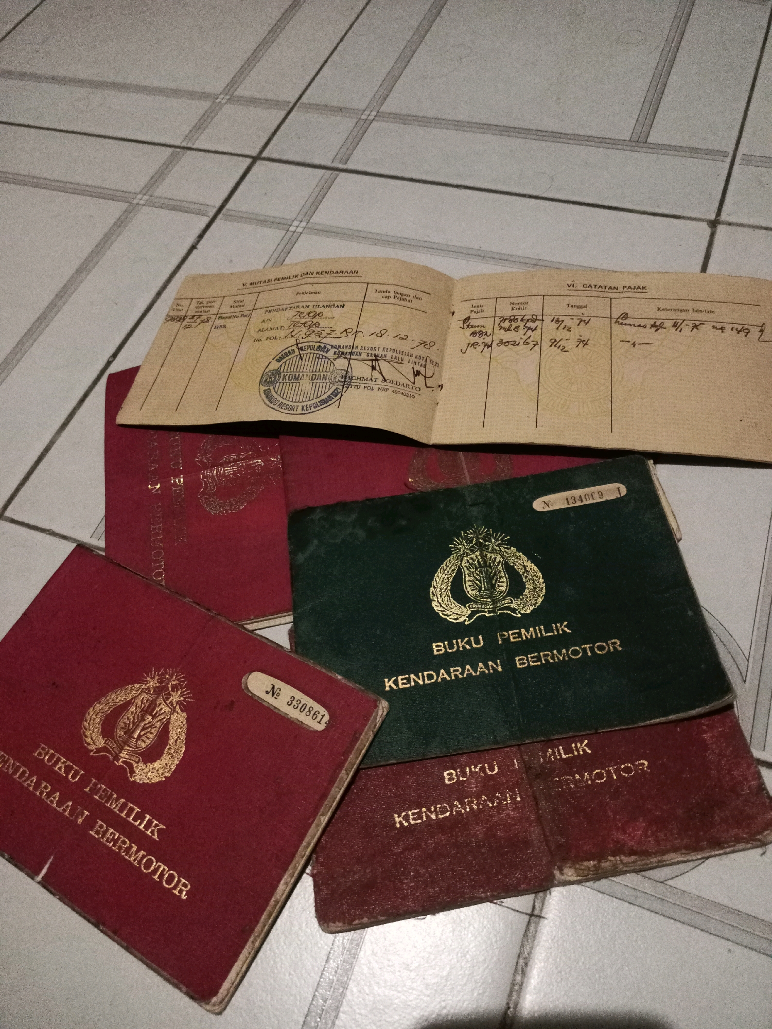 The image shows a collection of vehicle registration documents and related papers scattered on a tiled floor. The documents include several "BUKU PEMILIK KENDARAAN BERMOTOR" (Vehicle Ownership Book) in red and green covers, some with gold lettering and emblems. One of these red books has a sticker with the number "№ 3308614". There is also an open document with sections for "V. MUTASI PEMILIK DAN KENDARAAN" (VI. Vehicle and Owner Mutations) and "VI. CATATAN PAJAK" (VI. Tax Records), containing handwritten entries. The tiled floor appears to be an indoor surface, possibly in a residential setting. The overall scene suggests a disorganized collection of official papers. There are no people visible in the image, nor any specific indicators of the city of Malang, Indonesia. The lighting is dim, suggesting it might be indoors with artificial light or taken at night.