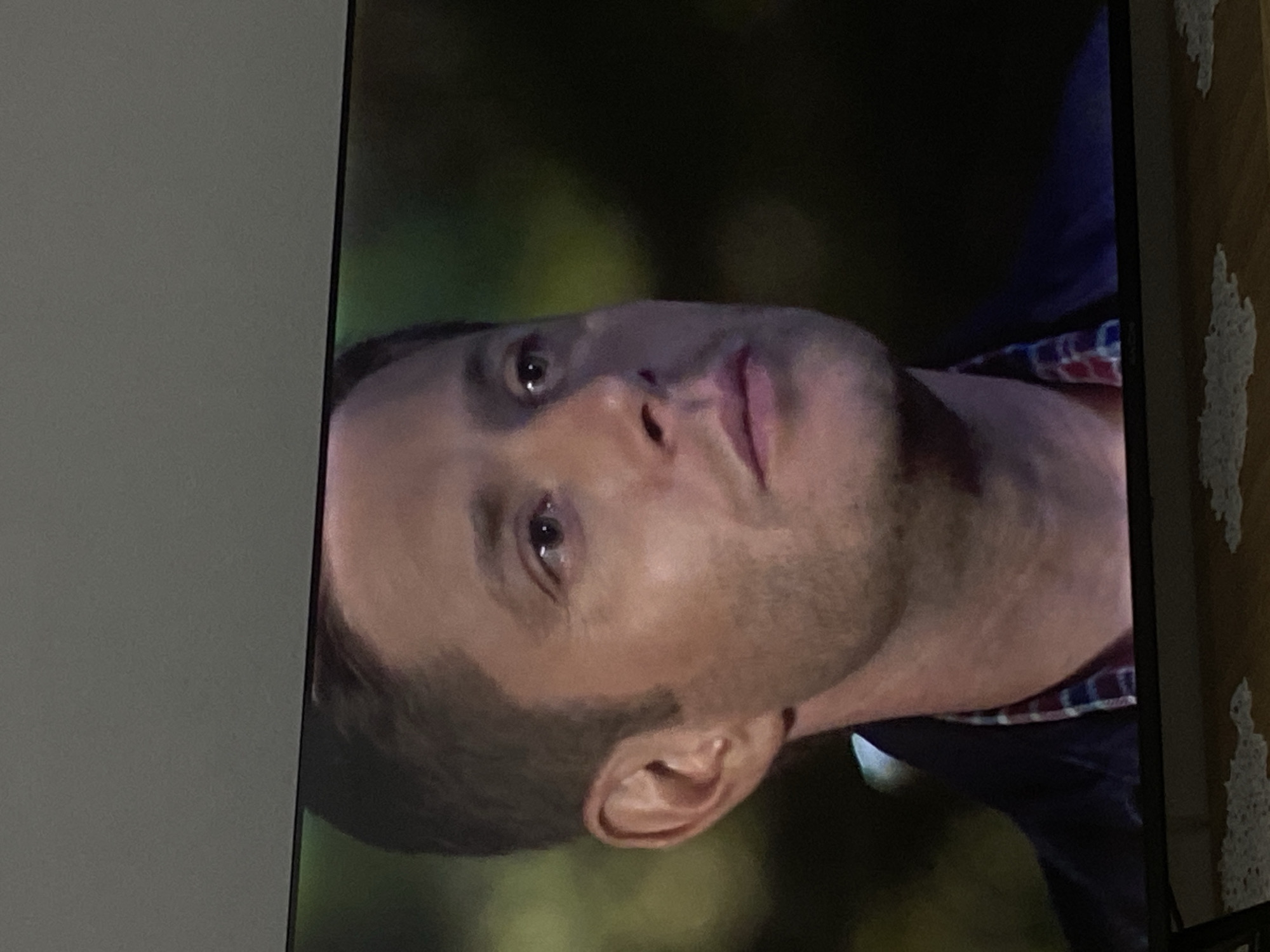 A close-up shot shows the face of a man, with his left side more visible. He has short brown hair, hazel eyes, and a slight stubble. His left ear is visible. He is wearing a dark jacket over a plaid shirt with red, blue, and white checks. The background is blurred, with hints of green and dark areas, suggesting an outdoor or dimly lit setting. The image appears to be a still from a television screen, with the bottom edge of the screen and a portion of a wooden surface with lace mats visible in the foreground. The location in the city of Konya, Turkey is not discernible from the visual information provided.