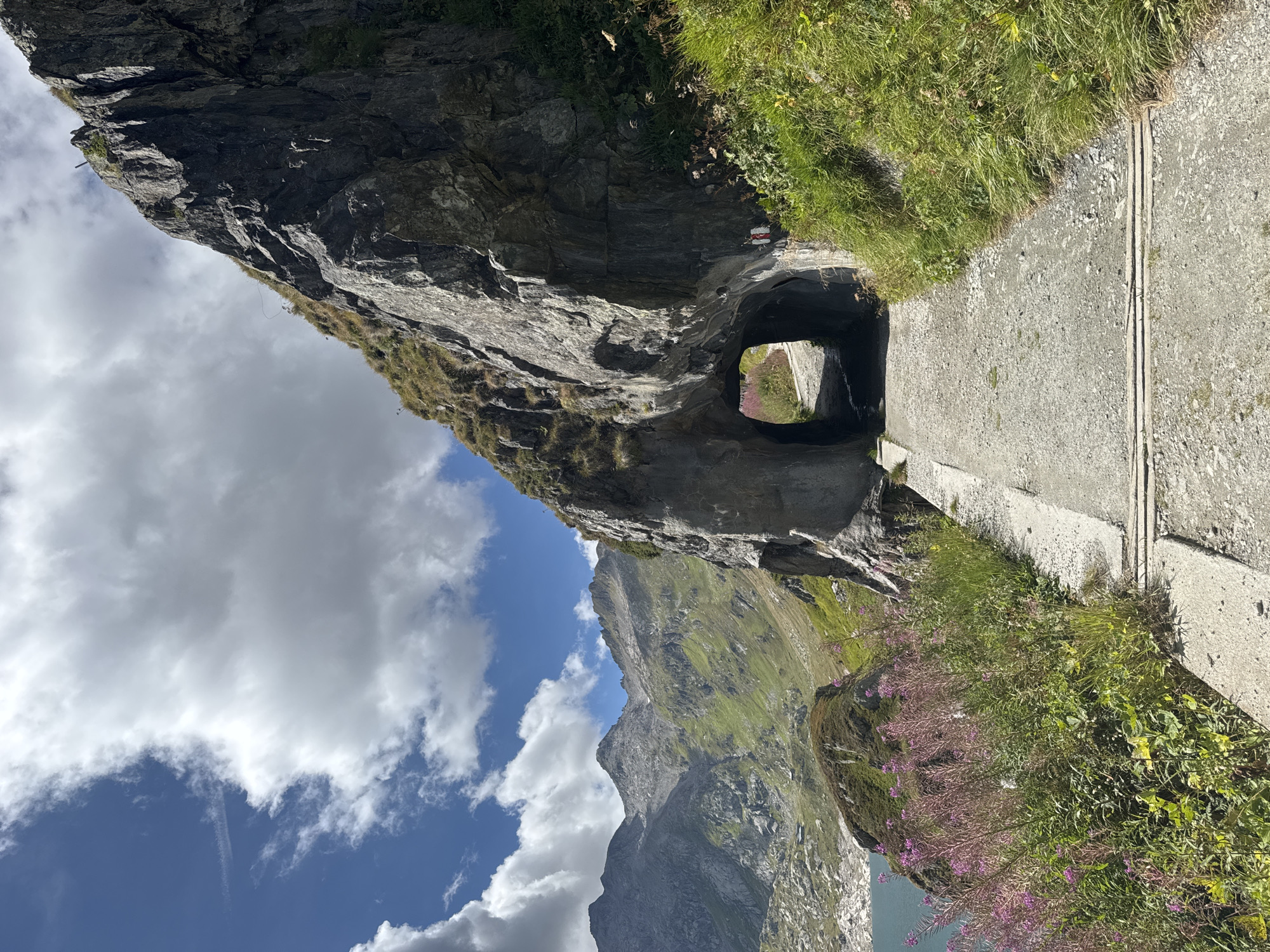 A gravel path leads through a natural rock archway in a mountainous landscape. The path is bordered by green grass and some purple wildflowers on the left. In the background, a serene lake is visible, reflecting the bright blue sky dotted with white clouds. Beyond the lake, rugged, green mountains rise majestically under the clear sky. A red and white trail marker is visible on the right side of the archway. The scene appears to be a scenic hiking trail in the Swiss Alps, likely near Airolo, Switzerland.