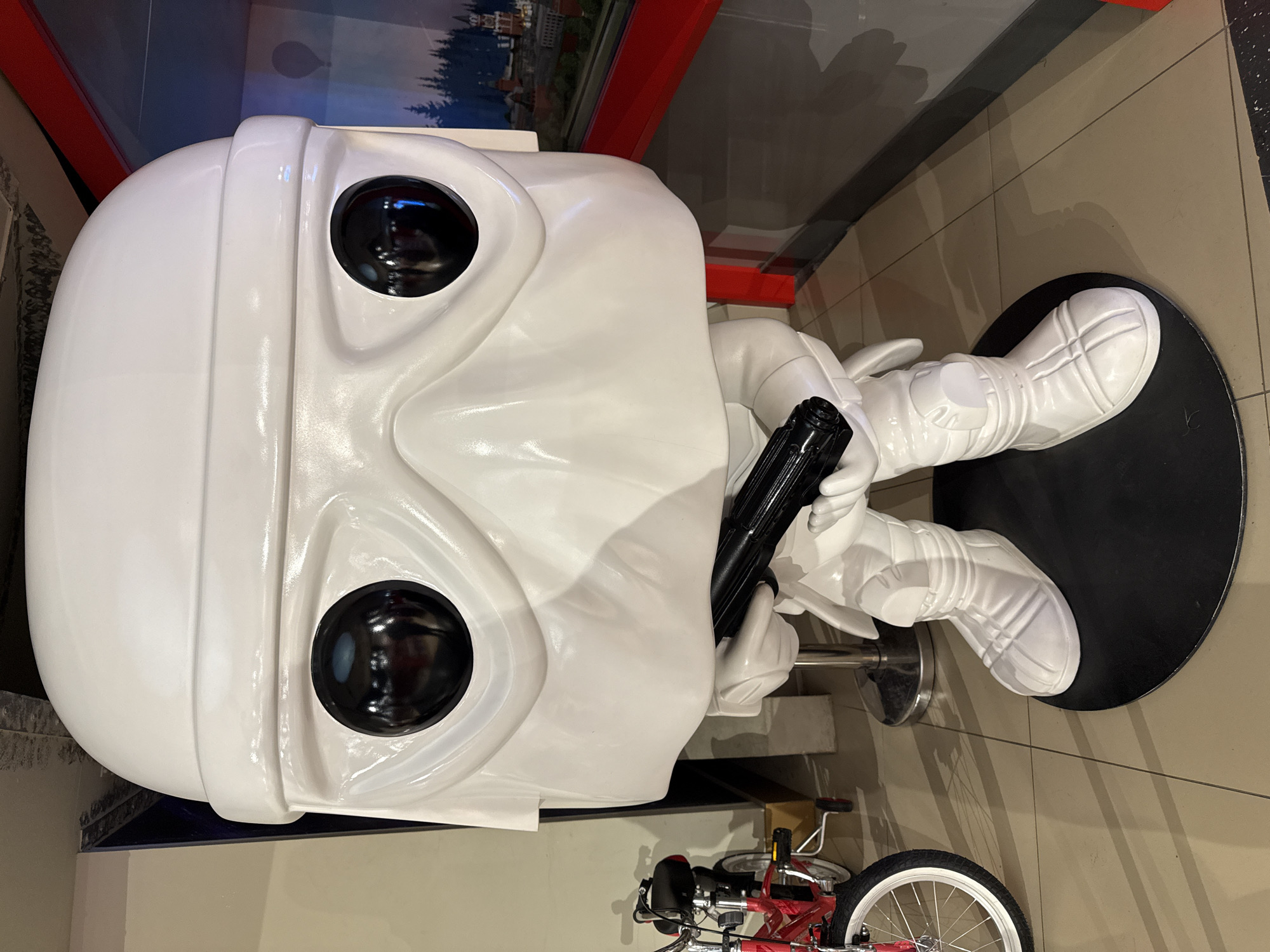 A large, white, Pop! Vinyl-style figure of a Star Wars stormtrooper is photographed indoors. The figure is holding a black blaster and sitting on a black circular base. The scene is in Unknown, Unknown. A red and white bicycle is visible in the background. The figure is glossy.