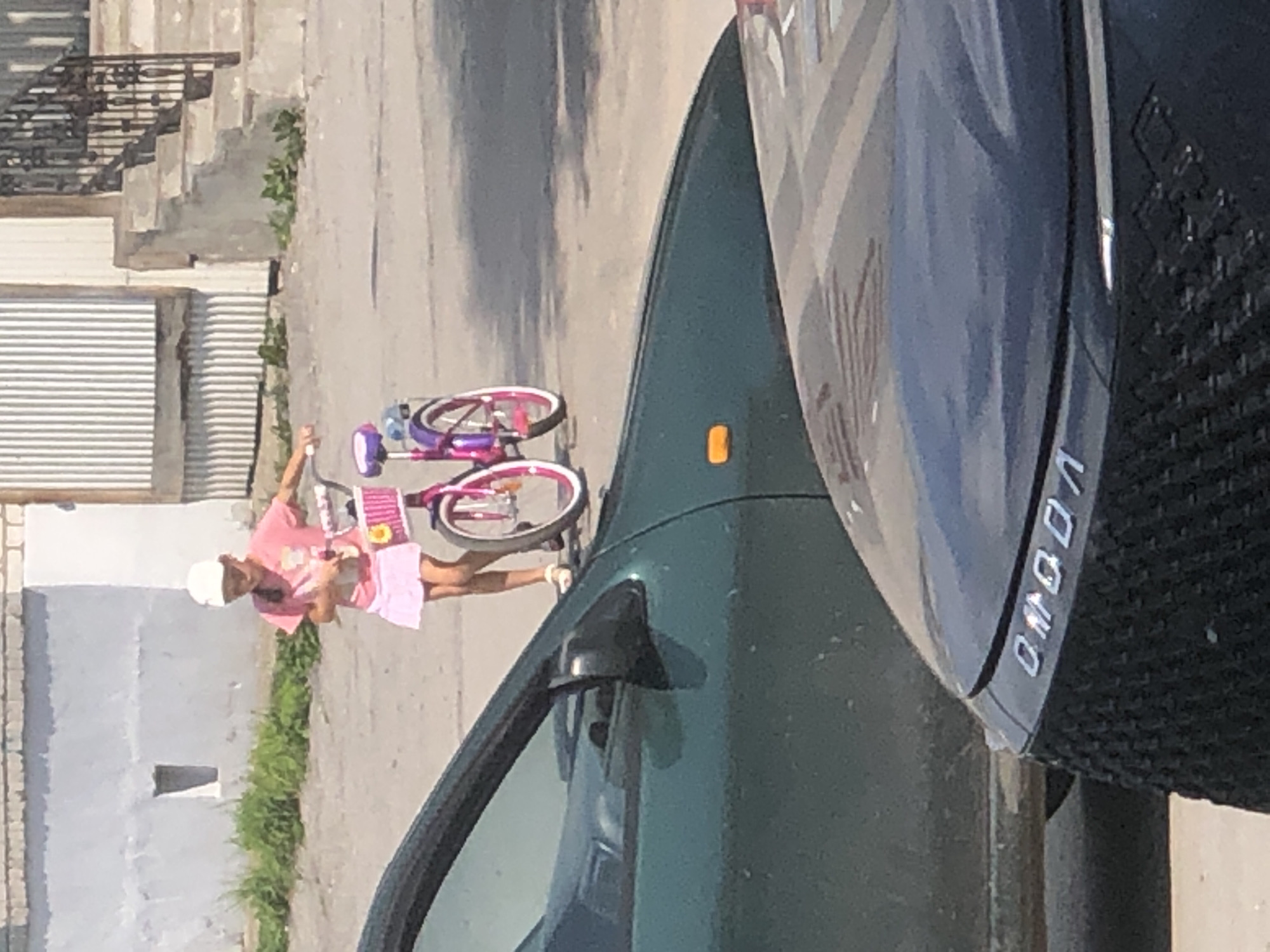 A young girl wearing a pink t-shirt, pink skirt, and a white cap is standing next to a pink and purple bicycle. She is holding onto the handlebars of the bicycle. The bicycle has a basket on the front with decorations. The girl appears to be looking down at something in her hands. The scene takes place on a paved area, possibly a street or sidewalk, with a green car in the foreground. In the background, there is a building with a corrugated metal siding and a set of stairs. The image is taken outdoors in daylight.
