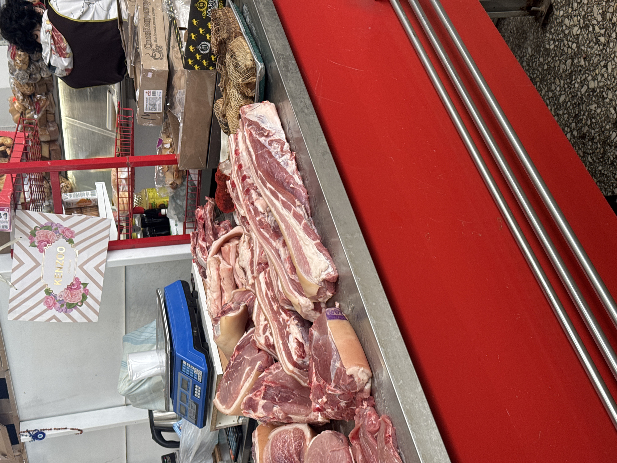 This image captures a vibrant, albeit slightly cluttered, indoor market or butcher shop, likely located in Chaikovsky, Russia. The primary focus is a long metal tray filled with various fresh cuts of raw meat, predominantly pork, including ribs and other lean and fatty portions. The meat rests against a bright red surface, where three parallel silver metal rods are also visible.
On the left side of the frame, a blue digital scale with multiple buttons and displays is present, poised for weighing products. Above it, a white shopping bag with "KENZOO" in gold lettering framed by a floral design hangs. Partially visible on the far left is a person, likely a vendor, wearing an apron with a floral pattern and a dark-colored garment.
The background reveals an array of packaged goods characteristic of a local market. Several brown cardboard boxes are hanging, one prominently displaying Cyrillic text: "СоюзКондитеръ" (SoyuzKonditer, or Union Confectioner) and the slogan "Мы объеденяем любовь" (We unite love), suggesting confectionery items. Another box shows "ЛОСИН...", likely part of a brand name. Other items include mesh bags containing unknown contents, and various other packages on shelves or hanging from red metal racks. The lighting is artificial, illuminating the scene brightly. The image conveys a busy, functional market environment.