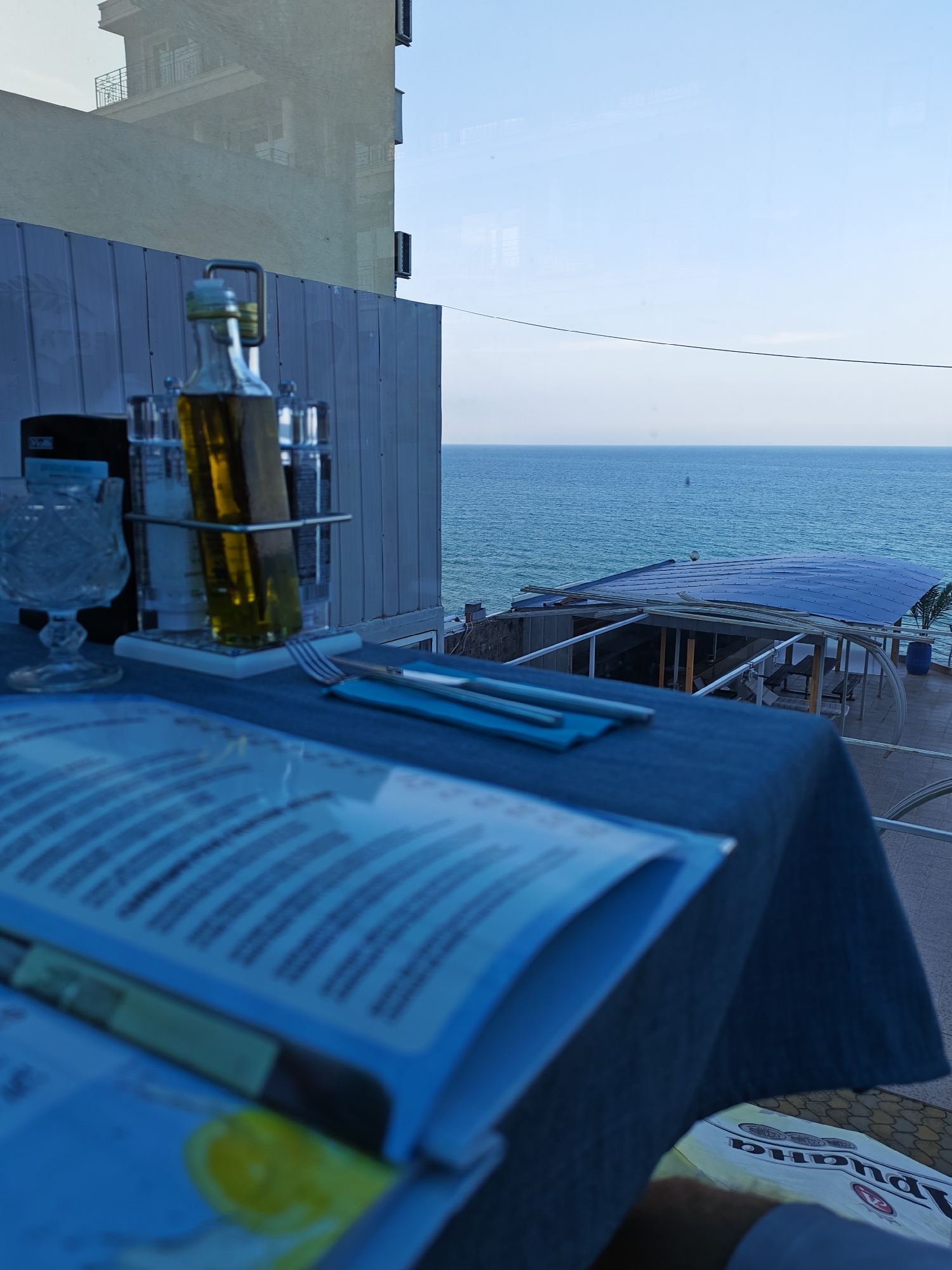 The image depicts a table set for dining at a restaurant with a view of the sea. The table is covered with a dark blue tablecloth, and on it are a menu, a crystal glass, a bottle of olive oil, salt and pepper shakers, and cutlery. In the background, the blue sea stretches to the horizon under a clear sky. A building is reflected in a window or glass surface on the left side of the image. To the right, an outdoor seating area with a blue canopy and tables is visible. The scene is set in Obzor, Bulgaria, a resort town on the Black Sea coast known for its beaches and seaside restaurants.