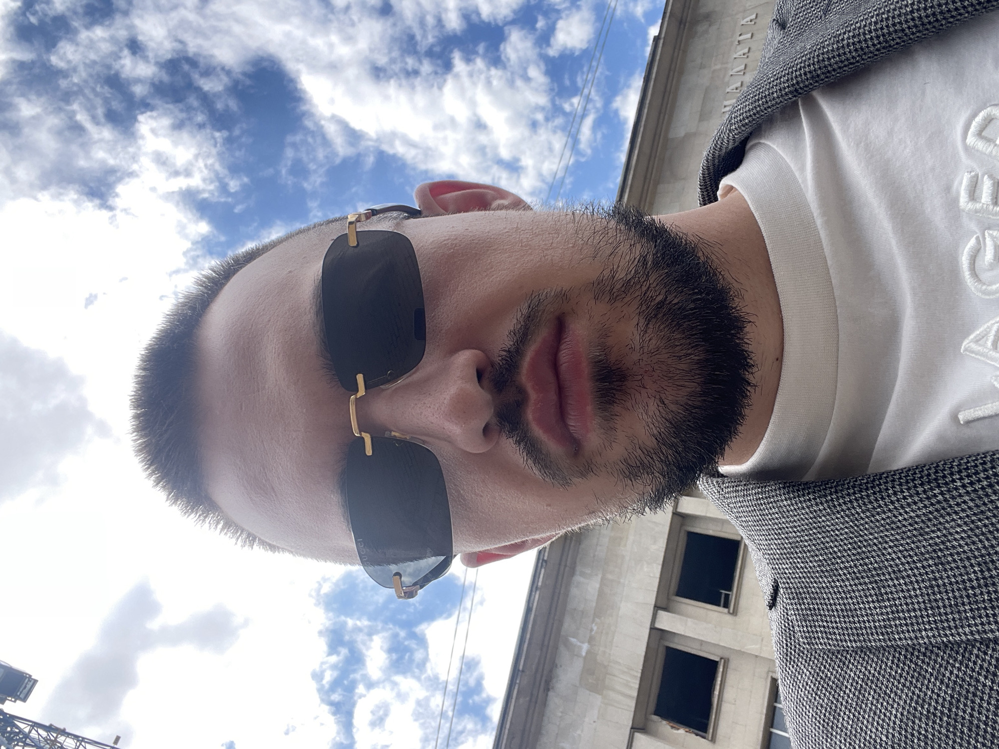 A low-angle photograph shows a man wearing dark sunglasses and a white t-shirt under a gray patterned blazer. The man has a short haircut and a dark beard. Above and to the left of the man is a blue sky with white clouds. To the right of the man is a building facade, partially visible, with several windows and lettering at the top. The lettering reads "НАДАТА". This suggests the location is Sofia, Bulgaria, and the building is the National Art Academy. A crane can be seen to the left of the man.