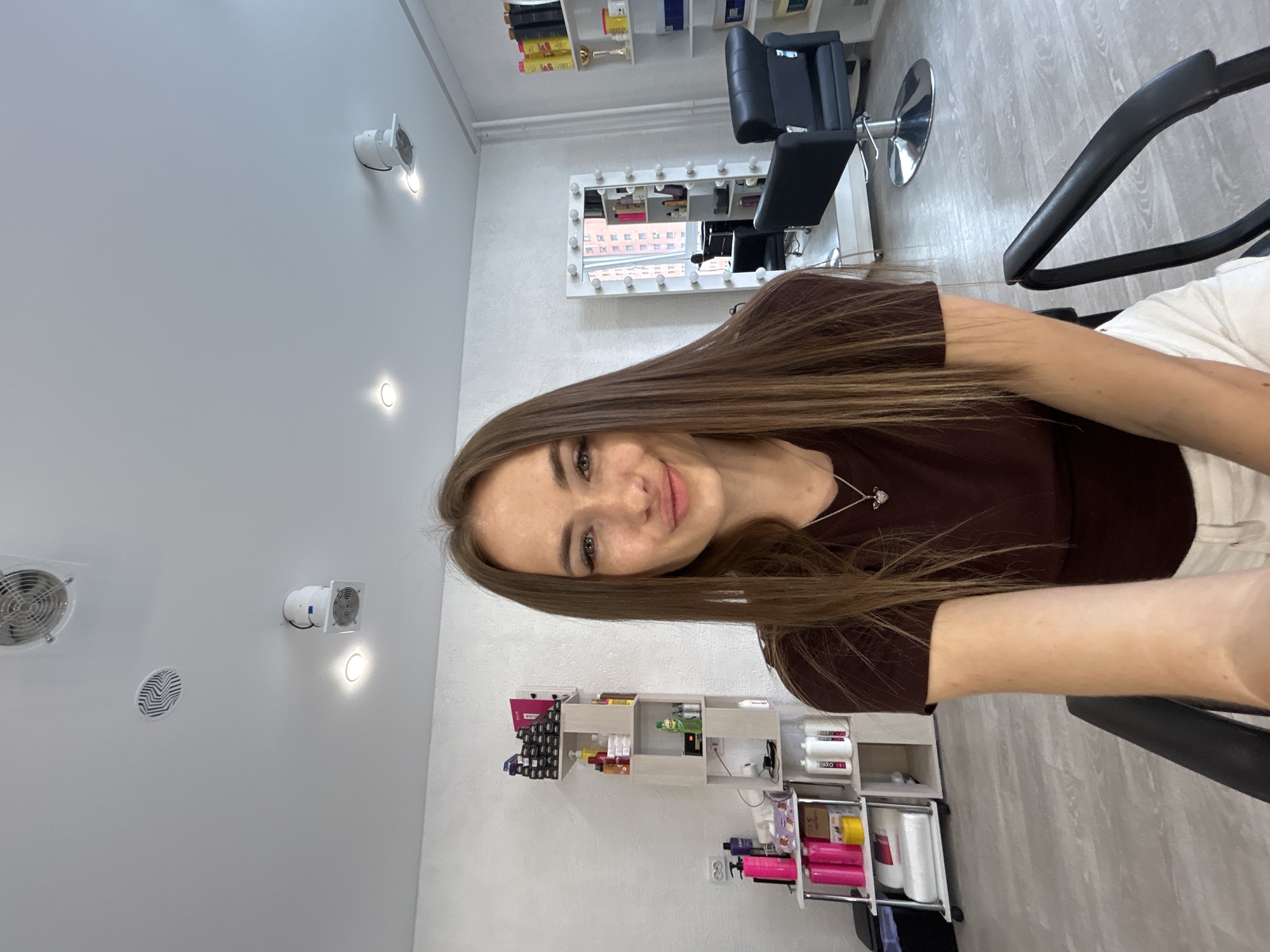 The image shows a young woman with long, brown hair, taking a selfie in what appears to be a beauty salon. She is wearing a dark brown top and white pants. Her expression is friendly, and she is smiling gently.
The background of the image reveals the salon's interior. There are shelves stocked with various beauty products, including bottles, jars, and tubes of different sizes and colors. A large mirror with lights around its frame is visible, suggesting it's a styling station. A black salon chair with a chrome base is also present. The walls are white, and the floor has a light-colored wood-like pattern. Several circular ventilation units are mounted on the wall.
The lighting in the image is bright, indicating it's likely daytime or the salon is well-lit. There are no strong shadows, suggesting ambient lighting. The overall atmosphere seems relaxed and professional. The selfie format and the woman's direct gaze at the camera indicate she is actively posing for the photograph.