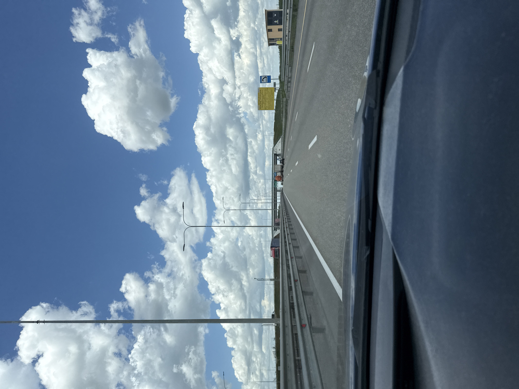 The image shows a view from inside a vehicle, likely a car, driving along a highway. The highway appears to be multi-lane and asphalt. The road is lined with streetlights. A guardrail is visible. In the distance, there are signs posted and a building. The sky is predominantly blue with numerous white, fluffy clouds. The exact location is Grushevskaya, Russia. There is no indication of human presence.
