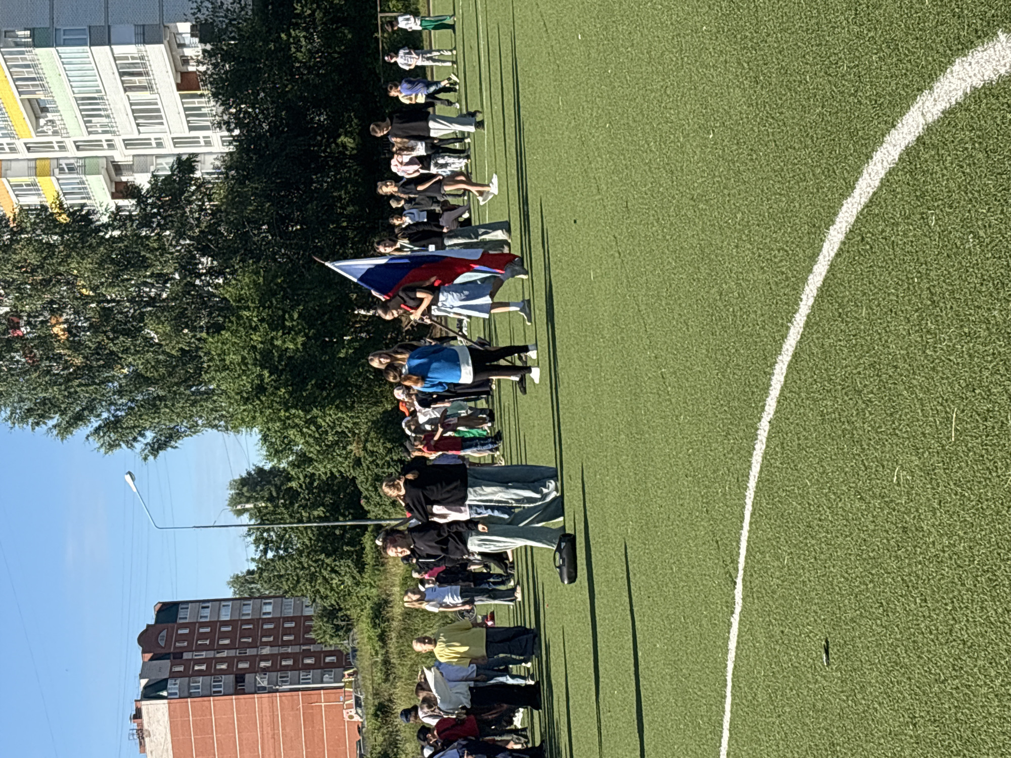 A group of young people, likely students, are gathered on a bright green artificial turf field. The field has a white curved line marking its boundary. In the background, a tall, modern building with balconies and white and colored facades is visible, along with trees and a blue sky. Some of the young people are holding a Russian flag. One individual in the foreground is wearing a black sweatshirt and light blue pants, while another next to them wears a blue hoodie and black leggings. To the left, a person in a yellow shirt and jeans stands out. A black electronic device, possibly a portable speaker, is on the ground in the middle of the field. The overall scene suggests an outdoor event or gathering, possibly a school activity.