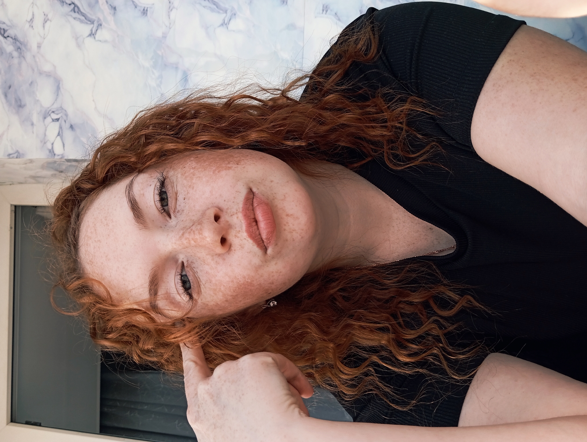A close-up, rotated portrait shows a young person with vibrant, curly red hair and numerous freckles scattered across their face. The person is lying down, looking upwards towards the camera with a subtle, contemplative expression. Their head is tilted, and one hand is raised, with a finger gently touching the hair or ear area.
They are wearing a ribbed black top, visible around the shoulders and neck. A delicate necklace with a small pendant is also visible on their chest. Their eyes are a light color, possibly blue or grey, and their lips are slightly parted.
The background primarily features a white and blue marbled pattern, which appears to be a wall or ceiling surface, indicating an indoor setting. Below the person's head, a darker, flat surface, possibly a window frame or dark furniture, is partially visible. The lighting is soft and even, suggesting it is not direct sunlight. There is no visible text in the image. The overall mood is calm and introspective.