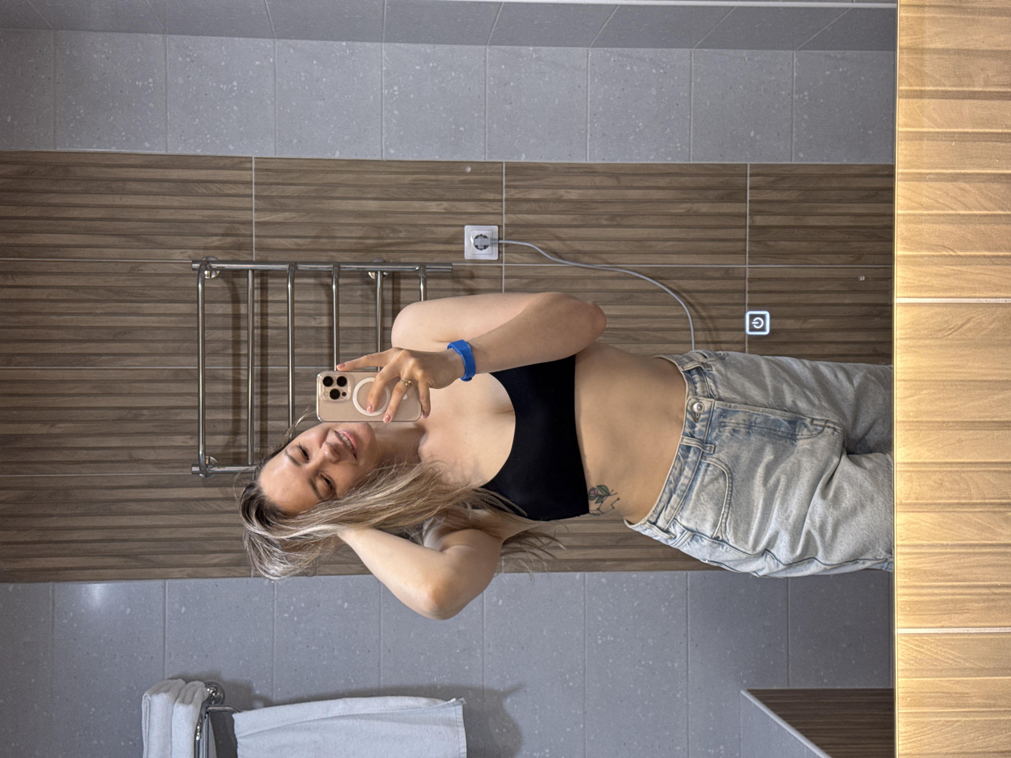This is a mirror selfie taken by a female in a modern bathroom in Nebug, Russia. The woman, appearing to be in her 20s or 30s with light-colored hair, is smiling and holding a gold-colored smartphone with a visible MagSafe ring in her left hand to capture her reflection. Her right hand is raised, touching her hair. She wears a black sports bra and light-wash denim shorts. A blue wristband is visible on her left wrist, and a small plant-design tattoo is on her right side, below her ribcage. The background reflects a bathroom interior featuring grey speckled wall tiles on the left and vertical brown wood-slat panels on the right. A silver-colored towel rack with white towels is mounted on the left wall, and a chrome-plated heated towel rail is against the wood-slat paneling. A white power outlet with a cord plugged in is visible on the wood-slat paneling. A glowing blue power symbol, indicating a touch-sensitive control, is present on the lower right portion of the mirror's surface. A light brown wooden or wood-patterned surface is reflected at the bottom edge of the mirror.