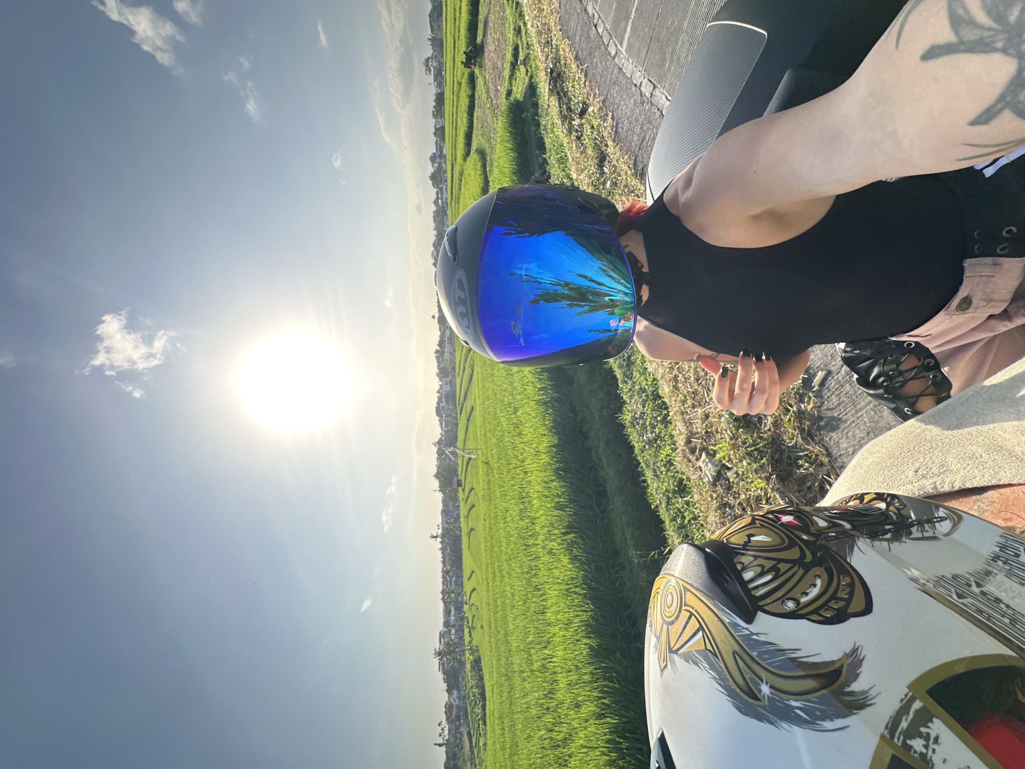 A person wearing a black sleeveless top and pink pants is seated on a motorcycle, taking a selfie. They have red hair and are wearing a black helmet with a blue reflective visor. In the foreground, another white helmet with gold and black markings is visible. The background depicts lush green rice paddies under a bright sun, with a few buildings and palm trees in the distance. The scene appears to be in North Kuta, Indonesia, given the characteristic landscape of rice fields and tropical vegetation. The sun is high in the sky, casting a bright light and creating a lens flare effect. The road is paved and curves to the right.