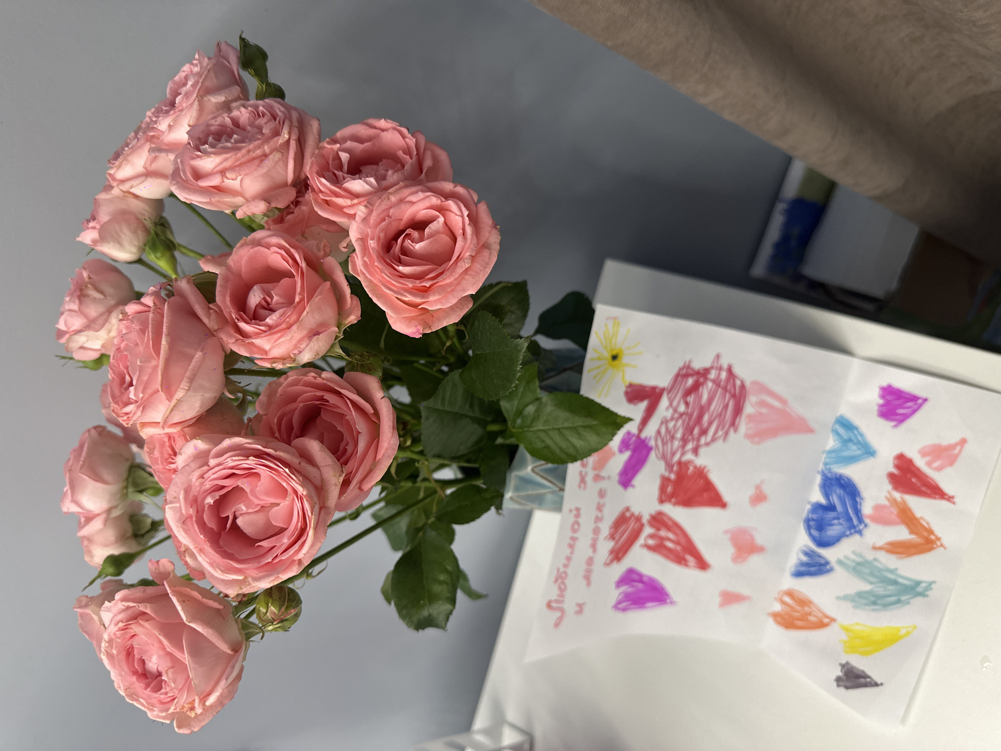 A top-down shot shows a bouquet of pink roses in a vase next to a child's drawing. The roses are tightly clustered, and their green leaves are visible. The drawing features colorful, abstract shapes, including hearts. Handwritten text in Russian is on the left side of the drawing. The background includes a light gray wall and a white table or surface. The precise location in Chaikovsky, Russia, cannot be determined from the visual information provided.