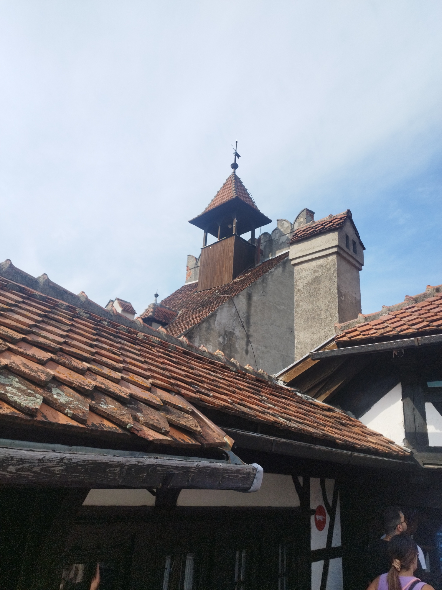 This media file captures an exterior view of historic architecture with a bright, clear sky overhead, suggesting fair weather and daytime. The scene is dominated by terracotta tiled roofs, some appearing weathered and moss-covered, indicative of age. In the background, a stone tower with a wooden structure at its peak rises above the rooftops, topped with a weather vane. Adjacent to it is a tall, square stone chimney with a layered top, also capped with tiles. The architecture suggests a European historical setting, possibly a castle or an old village.
In the foreground, the eaves of a building with dark wooden beams and white infill are visible, hinting at traditional construction. The lower portion of the image shows glimpses of people, with the back of a woman in a purple top and a man with glasses visible. They appear to be observing something, though their expressions are not discernible. A red and white circular sign is partially visible on the building's facade.
The overall impression is one of exploring a historical site. The clarity of the sky and the brightness of the light suggest it is a sunny day, likely late morning or early afternoon. The location context of Predeluț, Romania, further points to a site of historical and possibly touristic interest within the region. No discernible text is visible in the image beyond the small circular sign, which is not legible.