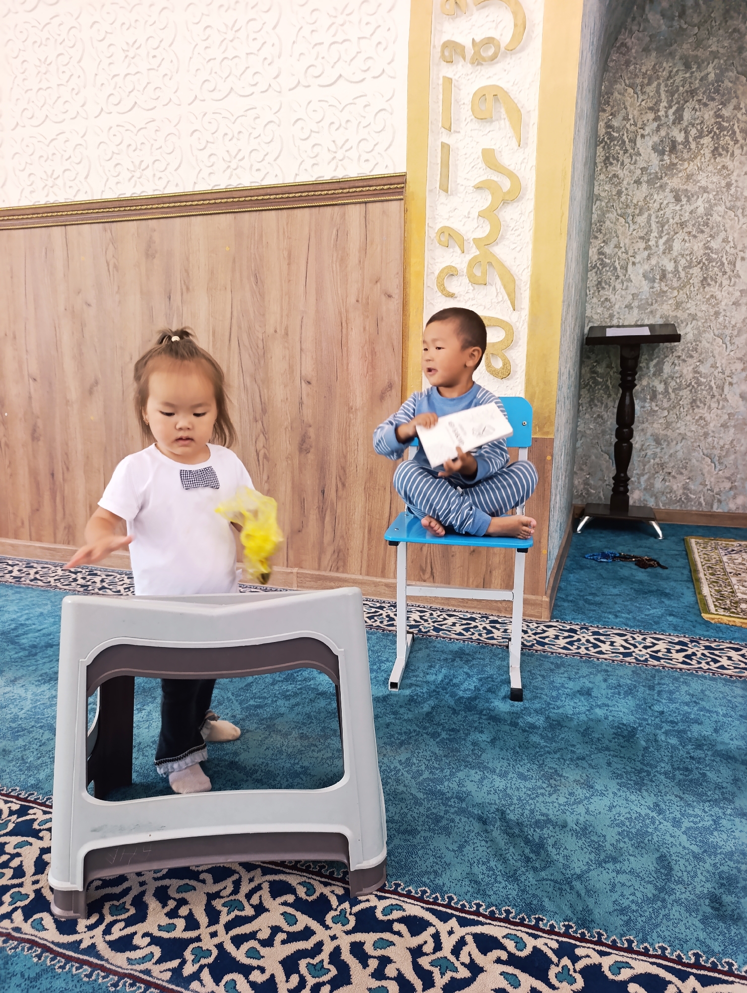 Two young children are inside what appears to be a mosque in Naryn City, Kyrgyzstan. A young girl with her hair in a ponytail stands in front of a white plastic stool, holding a yellow plastic bag. She is wearing a white t-shirt with a black and white bow tie. To her right, a young boy sits cross-legged on a blue chair, holding a white book. He is wearing blue and white striped pajamas. The room has a patterned wallpaper on the left wall, wooden paneling on the back wall, and a textured wall with gold Arabic calligraphy on the right. The floor is covered with a blue carpet with an intricate pattern. A dark wooden table with a black stand is visible on the right side of the image.