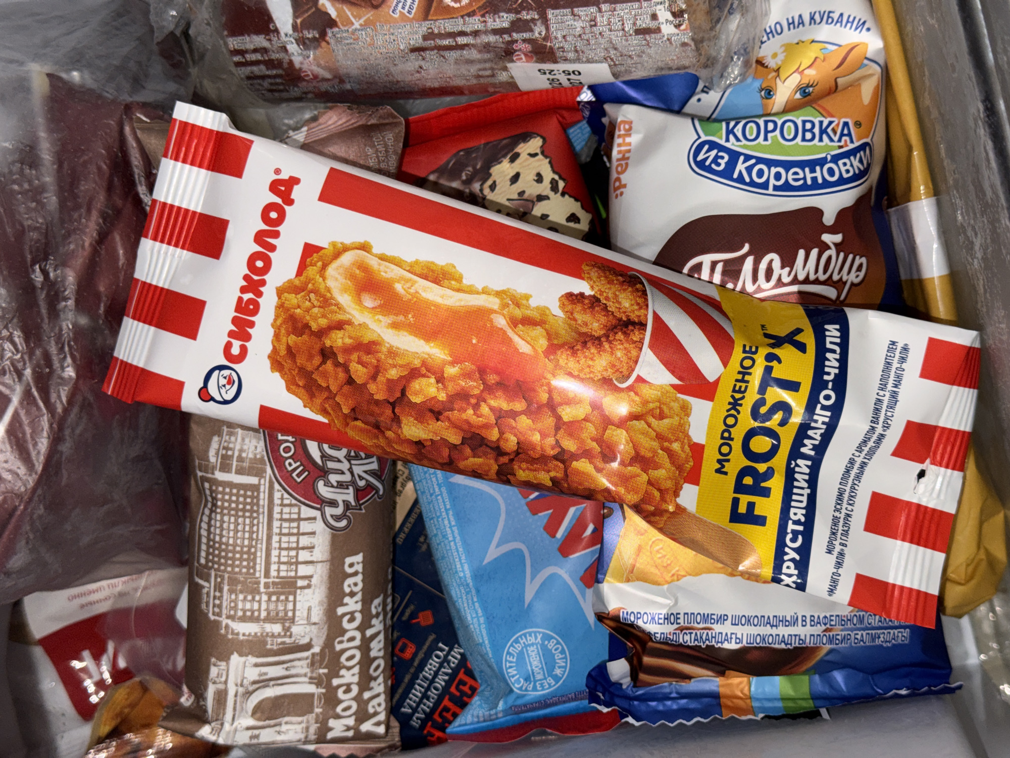 The image shows a close-up of several packaged ice cream bars in what appears to be a freezer. The ice cream bars are of various brands and flavors, with prominent Russian text on their wrappers. One prominent ice cream bar is red and white striped with an image of fried chicken on it, and the text "СИБХОЛОД" (Siberian Cold). Another ice cream bar has the text "Московская Лакомка" (Moscow Delicacy). There is also a blue wrapper with the text "РАСТИТЕЛЬНЫХ ЖИРОВ" (vegetable fats) and another white wrapper with "КОРОВКА" (Cow) and images of deer. The overall environment suggests a commercial setting, possibly a store freezer or a home freezer. There are no people visible in the image. The image does not provide any specific information about a location in the city of Moscow, Russia, beyond the brand name "Московская Лакомка".