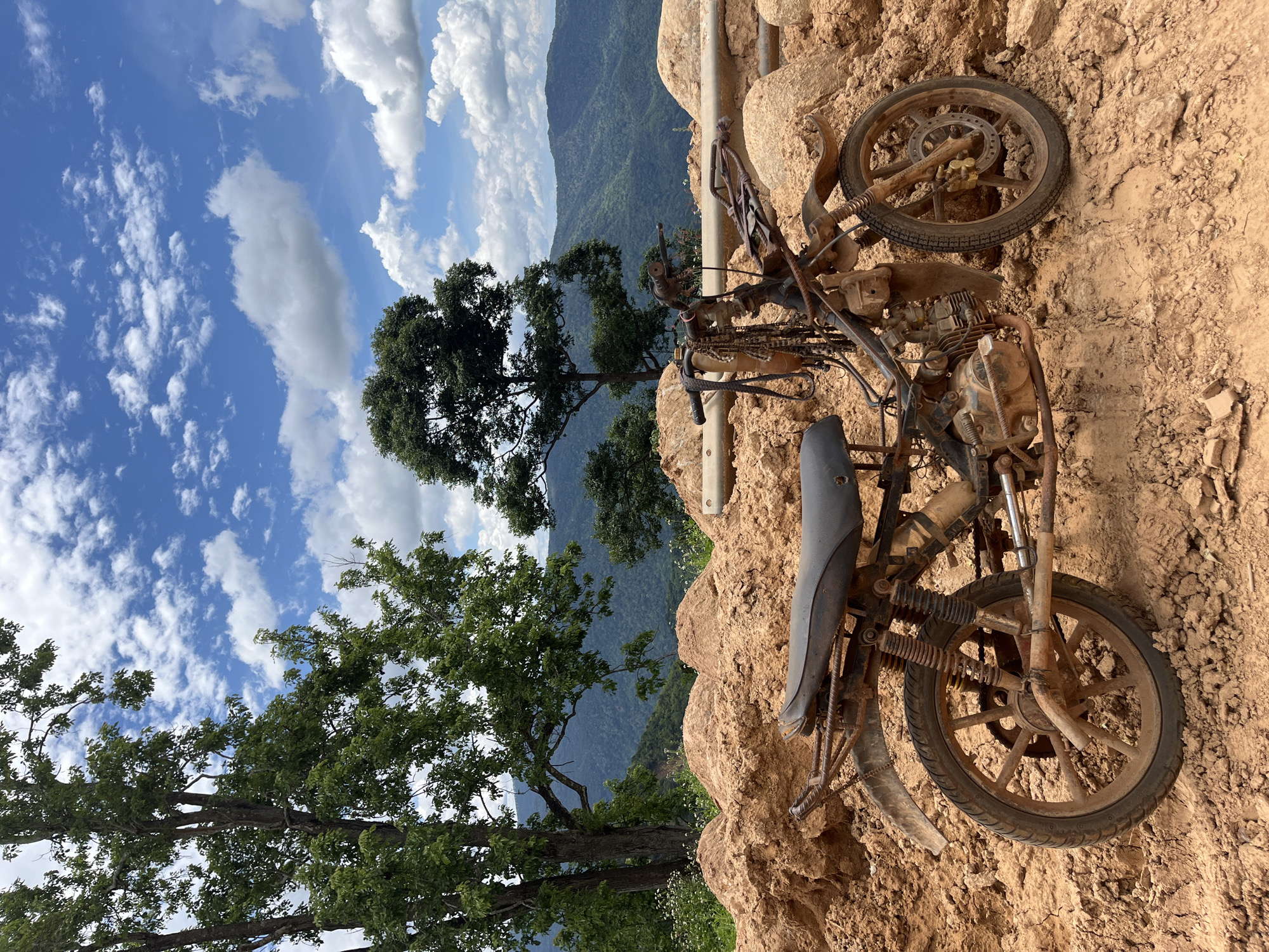 An abandoned, rusted motorcycle is parked on a dirt mound. The motorcycle is covered in dirt and debris, with its seat and handlebars visible. In the background, lush green trees and rolling hills create a scenic backdrop, under a bright blue sky with fluffy white clouds. The scene is set in a rural area, likely in the vicinity of Nao Se, Vietnam, given the landscape and vegetation. There are no people present in the image. The motorcycle appears to be a dirt bike or similar off-road model, its condition suggesting it has been exposed to the elements for an extended period. The surrounding terrain is rugged and uneven, composed primarily of reddish-brown soil and rocks, indicative of a construction site or a natural landslide area.