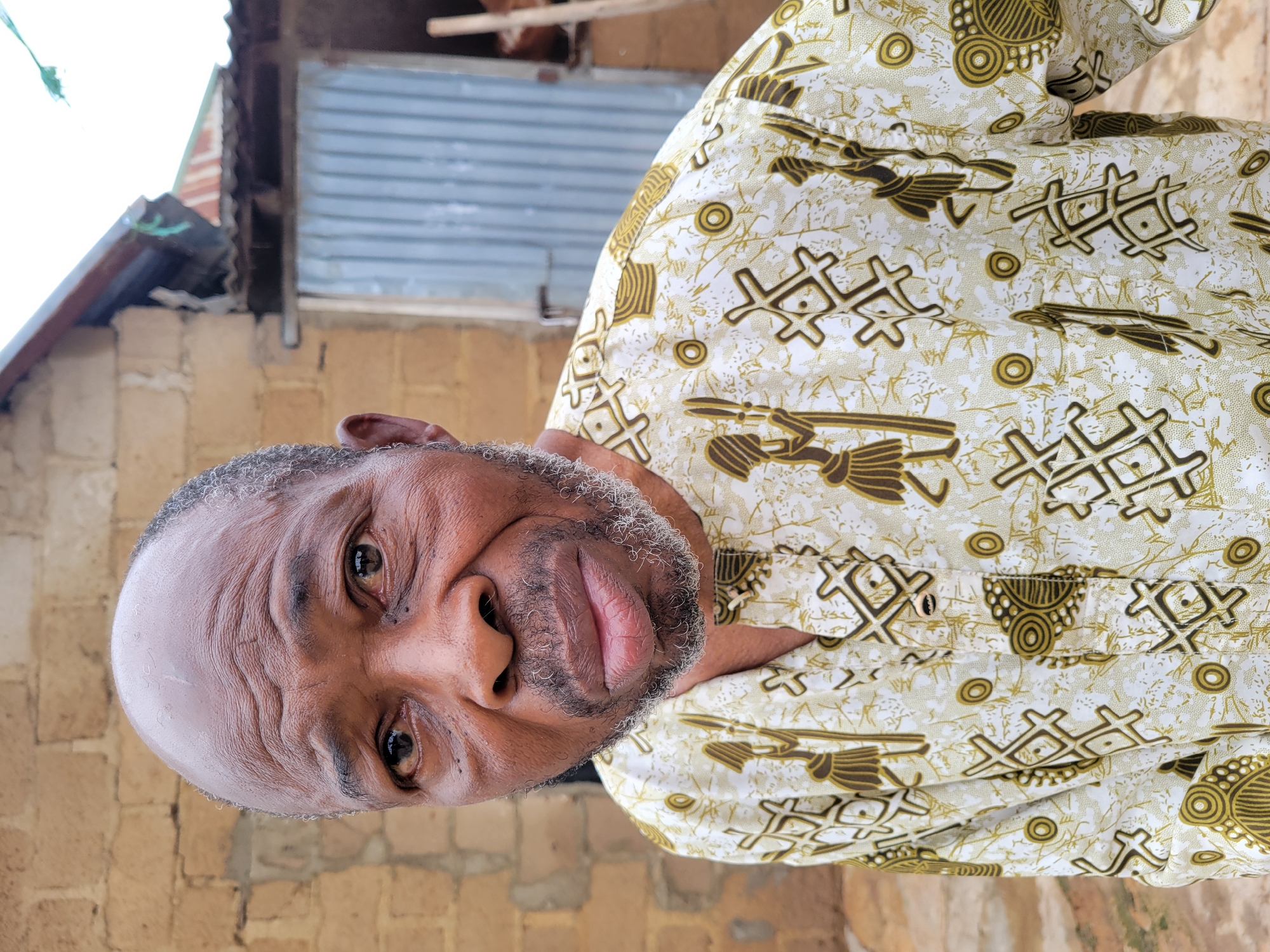 An older Black man is pictured from the chest up, looking directly at the camera with a neutral expression. He has a shaved head with graying hair around the sides and temples, and a graying beard and mustache. He is wearing a white long-sleeved shirt with a repeating pattern of brown figures that appear to be stylized people holding spears, interspersed with geometric shapes. The background is blurred, showing a brick wall on the left and a corrugated metal door and roof structure on the right. The environment appears to be outdoors, possibly in a rural or underdeveloped urban setting.