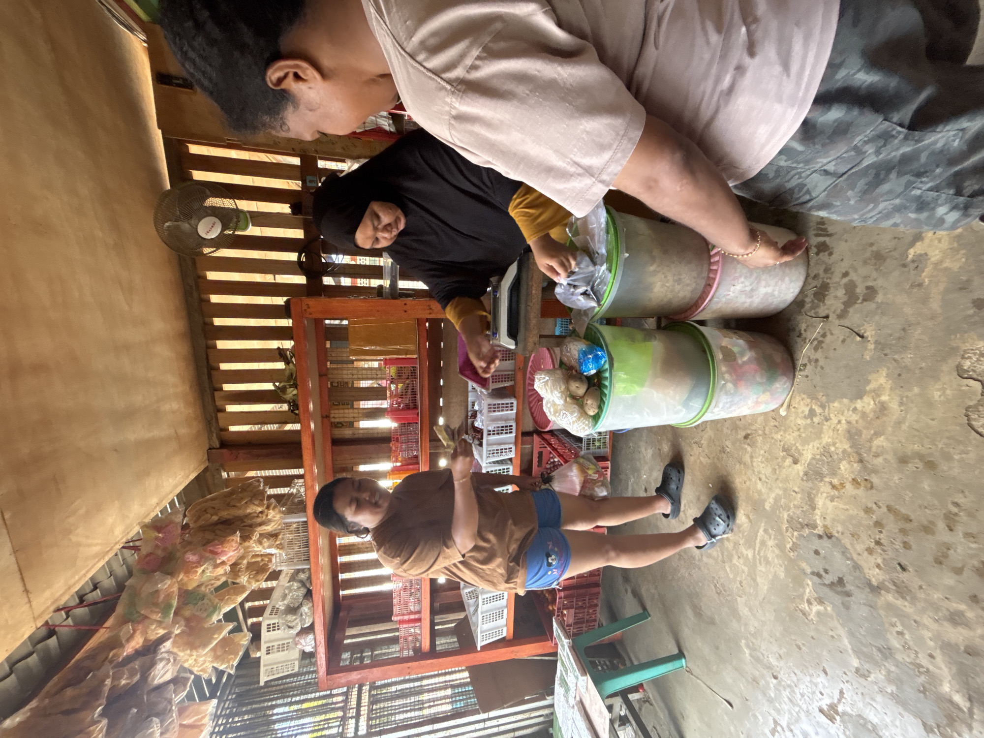 In a rural market stall in Merauke, Indonesia, a transaction is underway. A woman, wearing a brown t-shirt and blue shorts with Mickey Mouse motifs, stands holding cash. She is interacting with a vendor, who is wearing a black hijab and a yellow garment. The vendor is behind a counter, operating a small scale and handling bagged goods. The stall is open-air, with a rustic wooden roof and walls. Shelves behind the vendor are stocked with various items in baskets and containers. In the foreground, stacks of clear plastic containers hold food items, and a light green table with a stool is partially visible on the left. The ground is a dirt floor, wet and stained in places. Another person, viewed from behind and wearing a light-colored t-shirt and camouflage pants, stands between the viewer and the vendor, obscuring part of the scene. Hanging from the ceiling are multiple bags of dried snacks or food items.