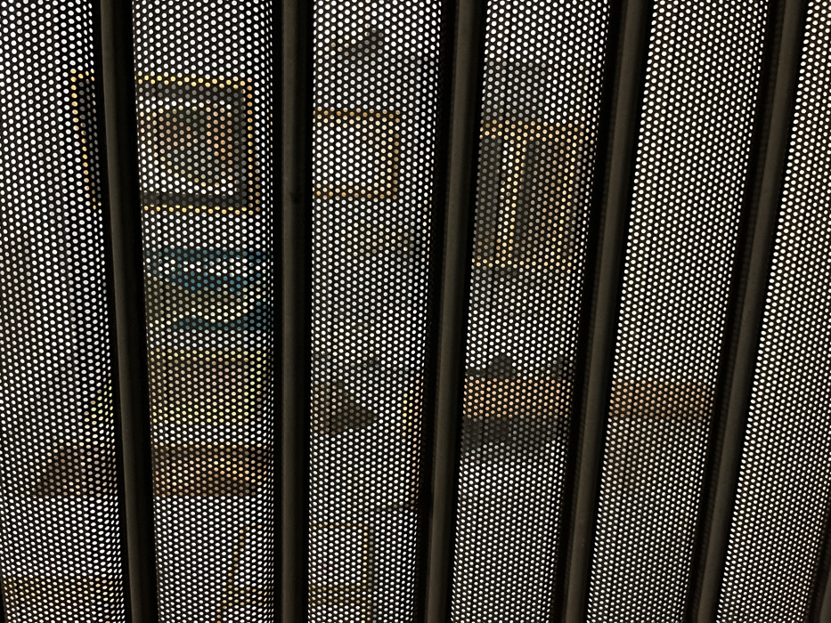The image presents a view through a perforated screen, which dominates the foreground and creates a highly pixelated and abstract perception of the background. The screen consists of dark, vertical bars separating panels covered in a uniform pattern of small, opaque white dots on a black surface, allowing light and blurred shapes to pass through.
Behind this visual filter, an interior space is dimly discernible. In the central-left area, a rectangular object is visible, appearing as a light-colored outline (possibly golden or yellowish) around a darker inner section, suggesting a framed display, a screen, or artwork. Directly below this rectangle, a distinct blue, somewhat rounded or rectangular shape can be observed. The surrounding background features further indistinct shapes and forms, primarily in warm, muted tones of yellow, brown, and hints of darker areas, indicative of furniture, walls, or other architectural elements within the room.
The scene depicts a static, obscured view of an interior, possibly a public space, gallery, or a room with modern architectural features. No specific activity is taking place, and no individuals are clearly visible. The lighting appears to be ambient indoor illumination. There is no visible text. While the image itself does not provide specific location cues, it is contextualized as being in Fiesole, Italy.