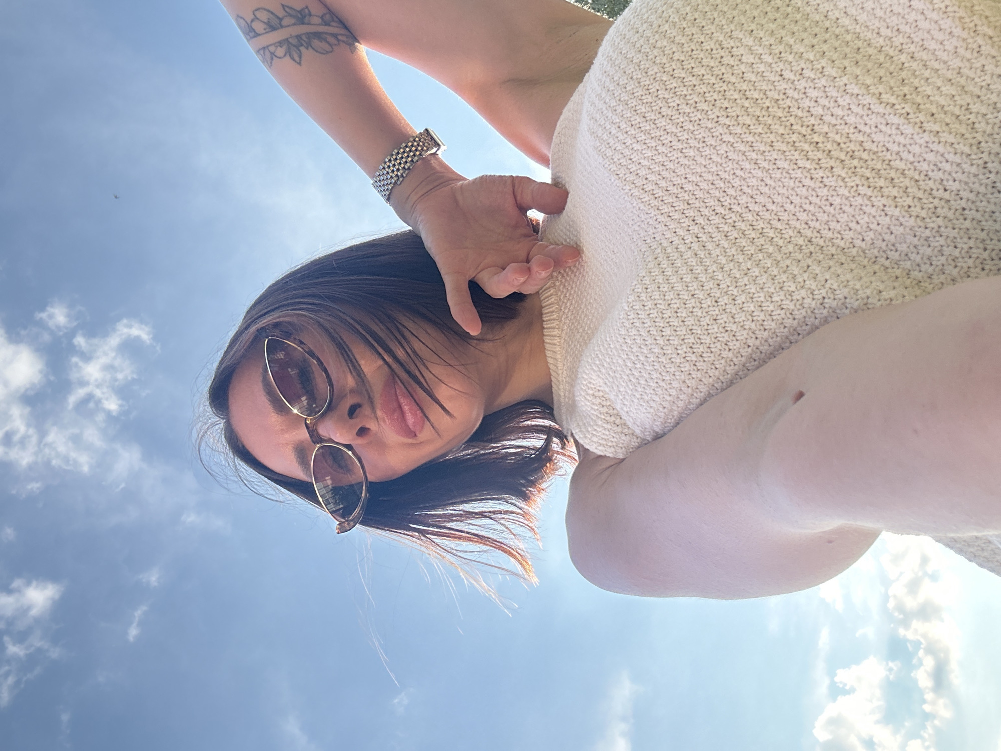 A low-angle shot captures a person from the shoulders up, facing the sky. The subject is wearing sunglasses with thin, gold-toned frames and a light-colored knitted top. Their short, dark hair is partially windswept. The sky is a bright blue with scattered white clouds. The person's left arm is raised, and they are wearing a silver-toned watch on their left wrist. A tattoo of floral designs is visible on their upper left arm. The overall scene is bright, with natural light illuminating the subject and the sky. There are no discernible landmarks or features that would place the scene in Votkinsk, Russia.