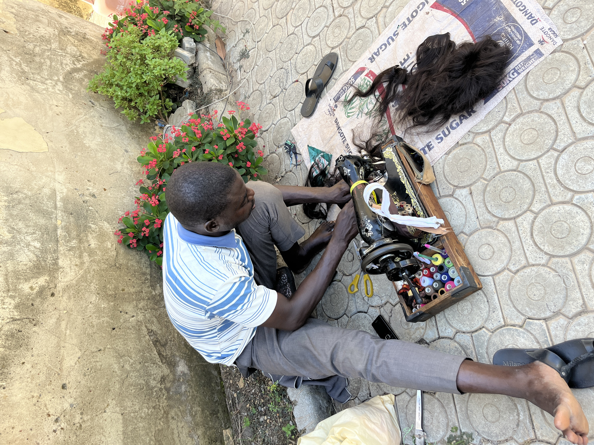 A man is operating a treadle sewing machine outdoors, likely in Abuja, Nigeria, as evidenced by the "DANGOTE SUGAR" bag serving as a workspace. He sits cross-legged on patterned concrete paving stones, wearing a blue and white striped shirt and gray trousers. A variety of threads are near the sewing machine. A clump of dark hair rests on the sugar bag. Nearby are a pair of black sandals, a pair of yellow scissors, and a black smartphone. The setting includes a concrete wall with flowering plants at the edge.