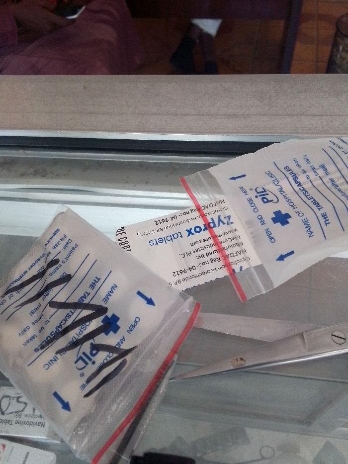 The image presents a close-up, downward-angled view of medical supplies arranged on a reflective surface, likely a countertop or display case. Three transparent plastic ziplock bags, designed for dispensing medication, are the primary focus.
The bag in the foreground, positioned towards the lower left, contains multiple white capsules. It features a red zipper closure and blue printed text, including "OPEN AND CLOSE HERE" with a directional arrow, a blue cross logo, "PID," and fields for "NAME OF HOSPITAL/CLINIC" and "THE TABLETS/CAPSULES." Black marker scribbles partially obscure some of this text. Another similar bag is partially visible above and to the right of the foreground bag.
A third bag, prominently displaying a white label, is positioned centrally. This label identifies its contents as "Zyprox tablets," specifying "Ciprofloxacin Hydrochloride B.P. 500mg." The label also details "Manufactured by: McCurre Industries PLC" and displays a "NAFDAC Reg no:- 04-9612," indicating the product is regulated by Nigeria's National Agency for Food and Drug Administration and Control.
To the right of the medication bags, a pair of silver-toned scissors with a gold-colored pivot screw is visible, partially obscured underneath the bags. The background above the counter features a beige, textured surface. Above this, a blurred reflection shows a person in a maroon or purple garment holding a white object, and a darker, indistinct shape, suggesting an indoor environment such as a pharmacy or clinic. The NAFDAC registration number confirms the location as Nigeria. The image contains no visual information to specifically identify the location within the city of Muhammadu Dikko Stadium.